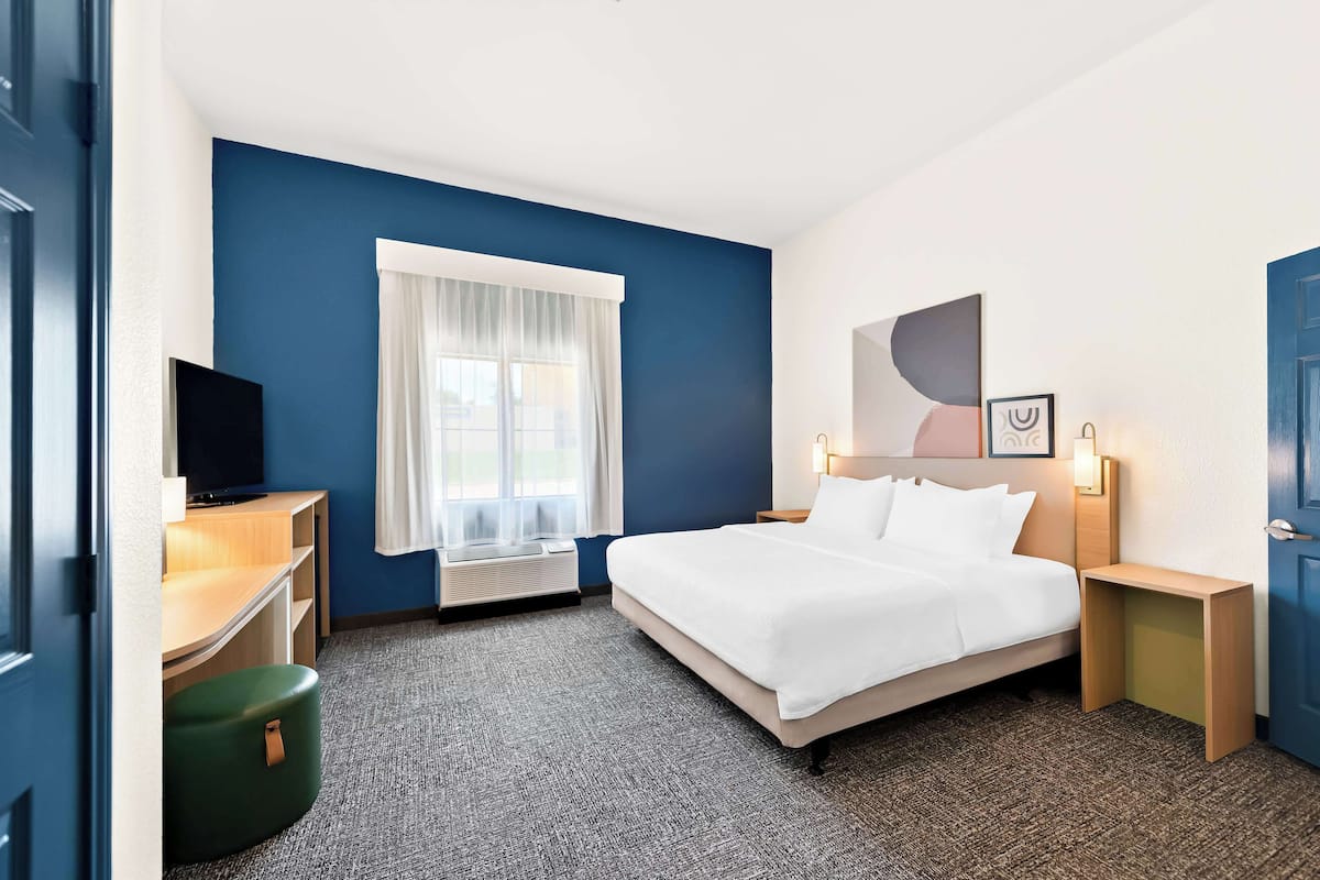 A hotel room with a bed, a desk with a computer, a window with curtains, and a wall-mounted TV.