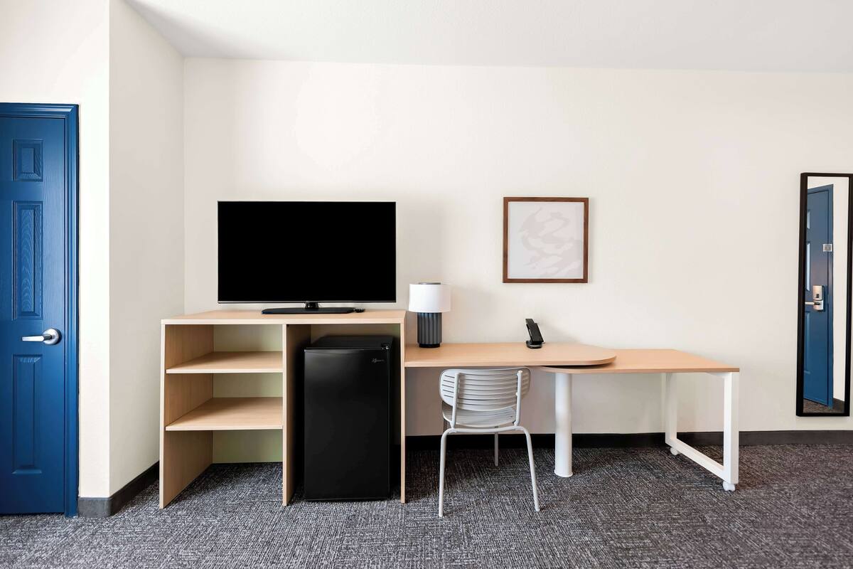 A compact hotel room with a single bed, a desk with a computer, a small refrigerator, a chair, and a television.