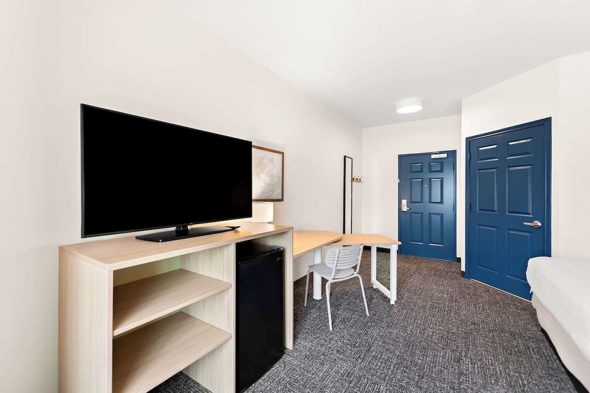 A hotel room with a TV on a wooden stand, a desk with a chair, a small fridge, and two blue doors.