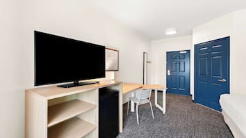 A hotel room with a TV on a wooden stand, a desk with a chair, a small fridge, and two blue doors.
