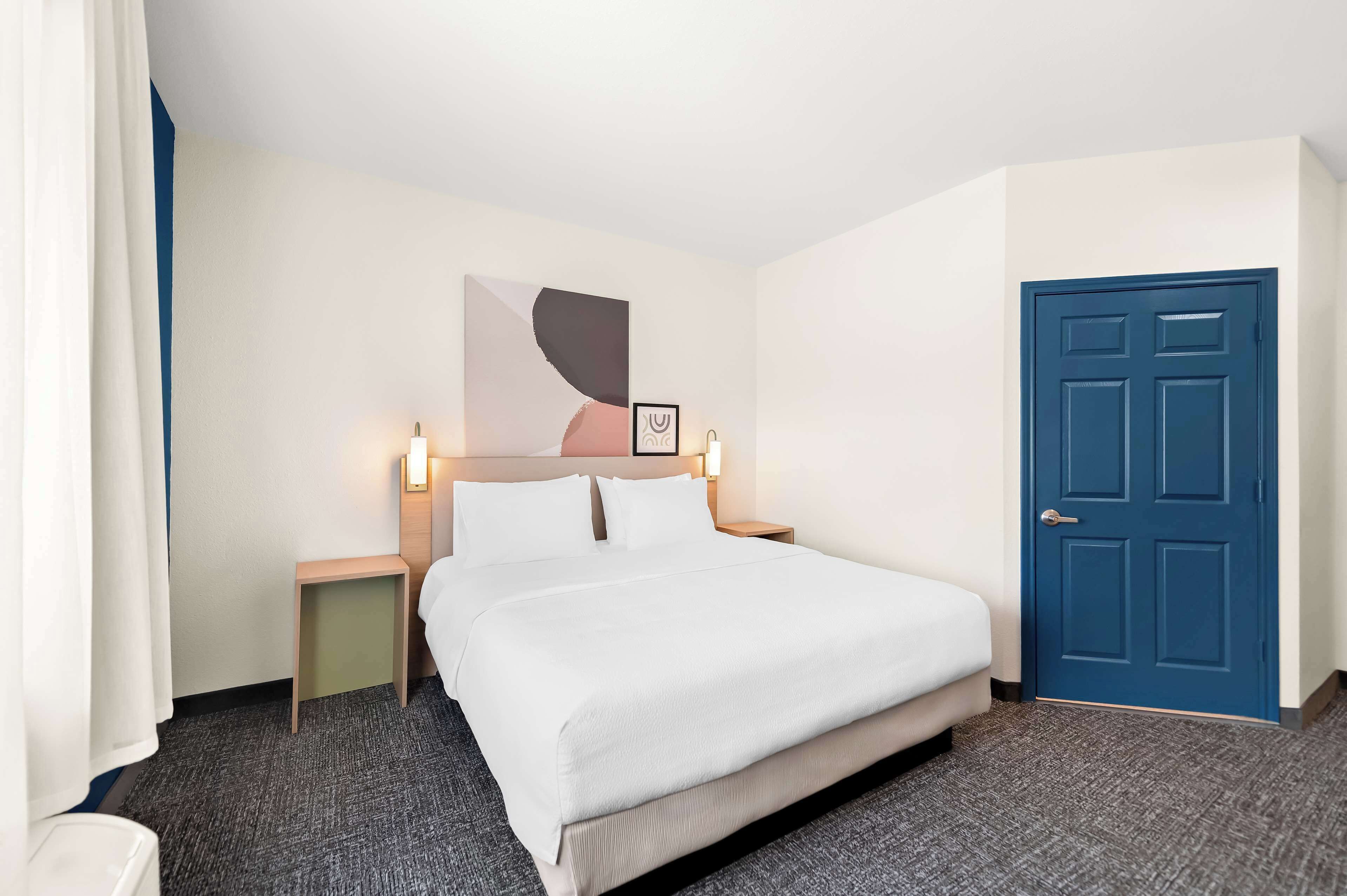 Une chambre d’hôtel avec un lit, une porte bleue, une table de chevet avec une lampe et un tableau encadré au mur.
