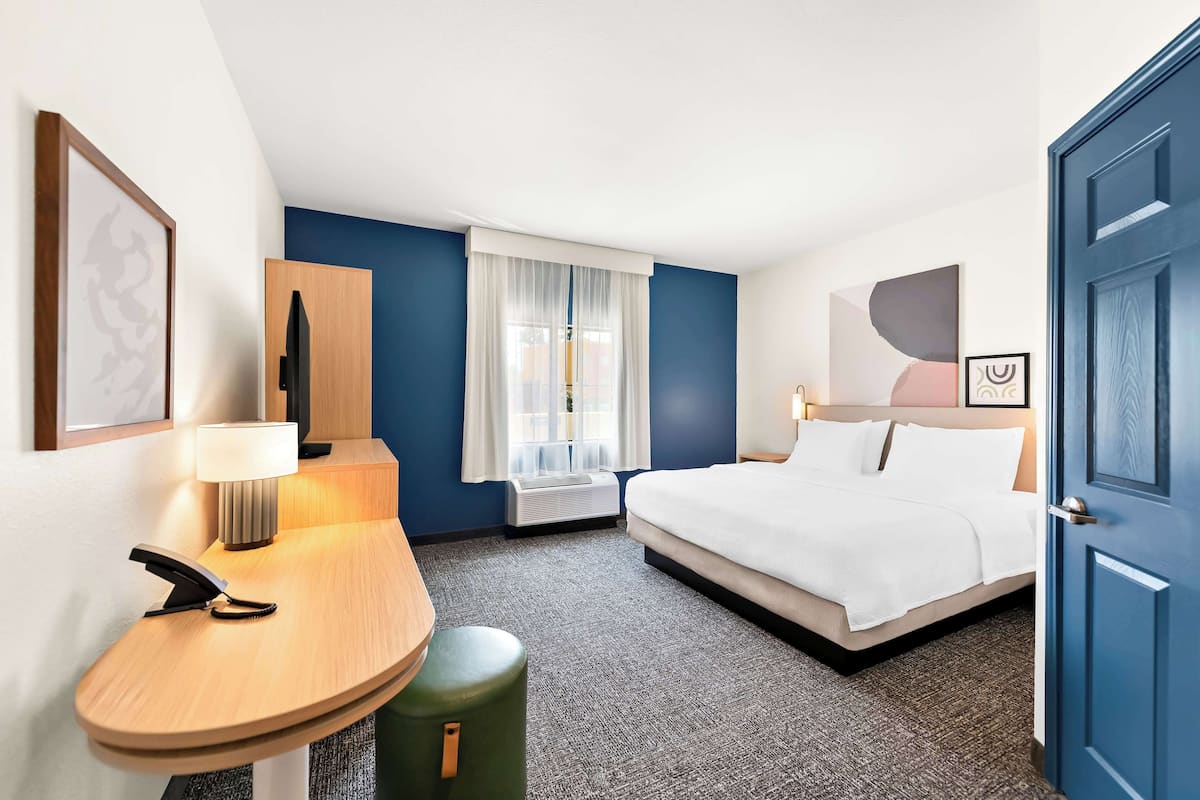 A hotel room with a bed, a desk with a telephone, a lamp, a window with curtains, and a door.