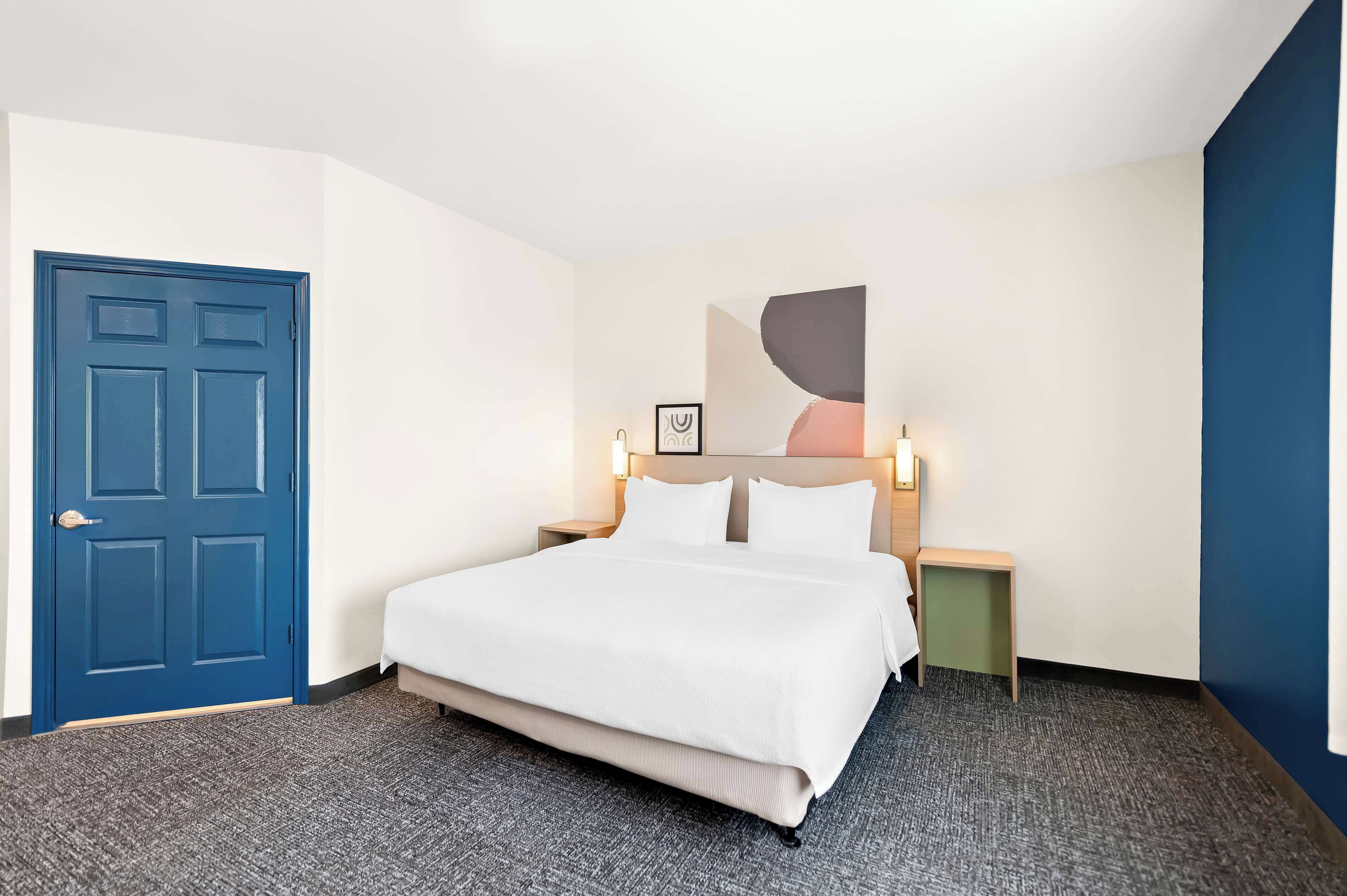 Una habitación de hotel con una cama grande, una puerta azul, una mesita de noche con lámpara y una obra de arte colgada en la pared.