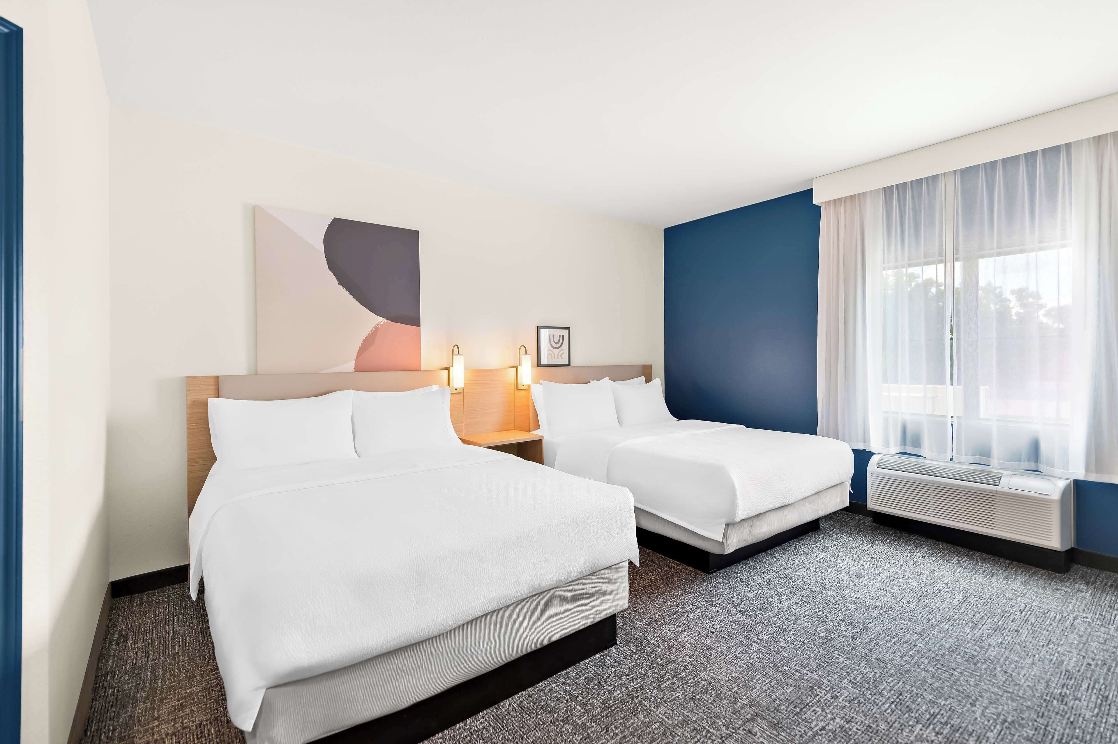 Een hotelkamer met twee bedden, een groot raam met dunne gordijnen, een luchtverkoeler aan de muur en een ingelijst schilderij boven de bedden.