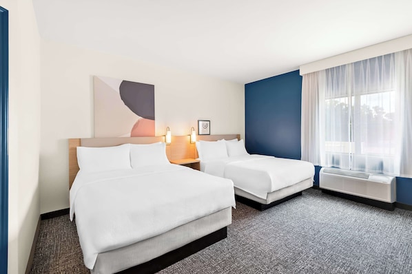 A hotel room with two beds, a large window with sheer curtains, a wall-mounted air conditioner, and a framed picture above the beds.