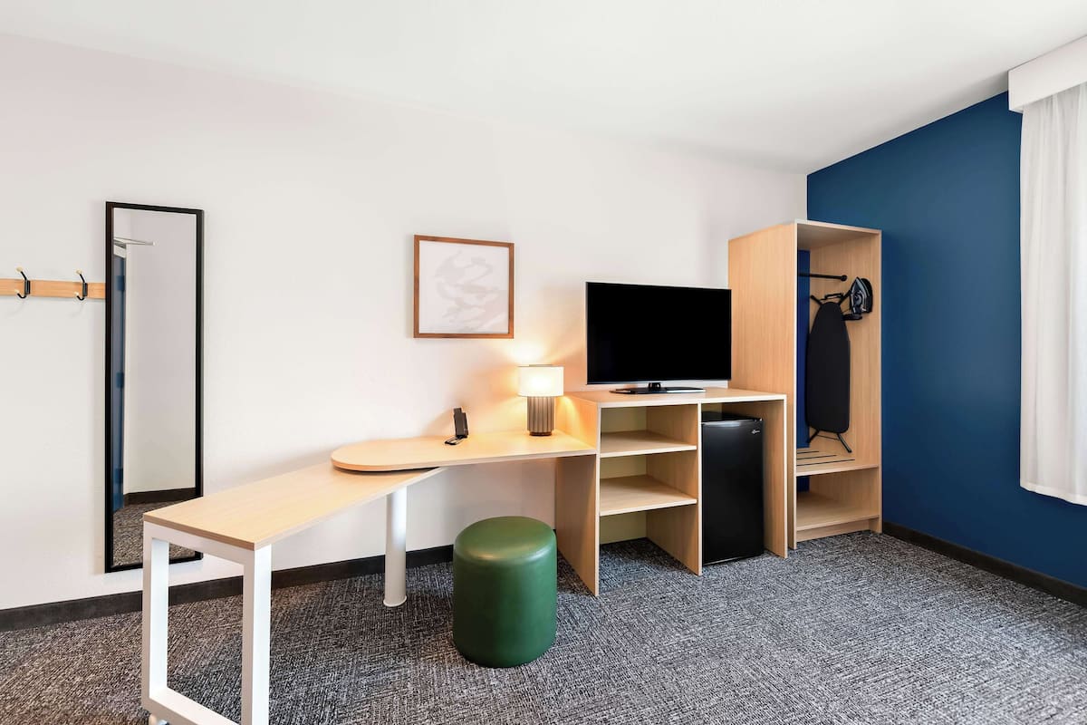 A hotel room with a desk, a television, a lamp, a green stool, a coat rack, and a wardrobe.
