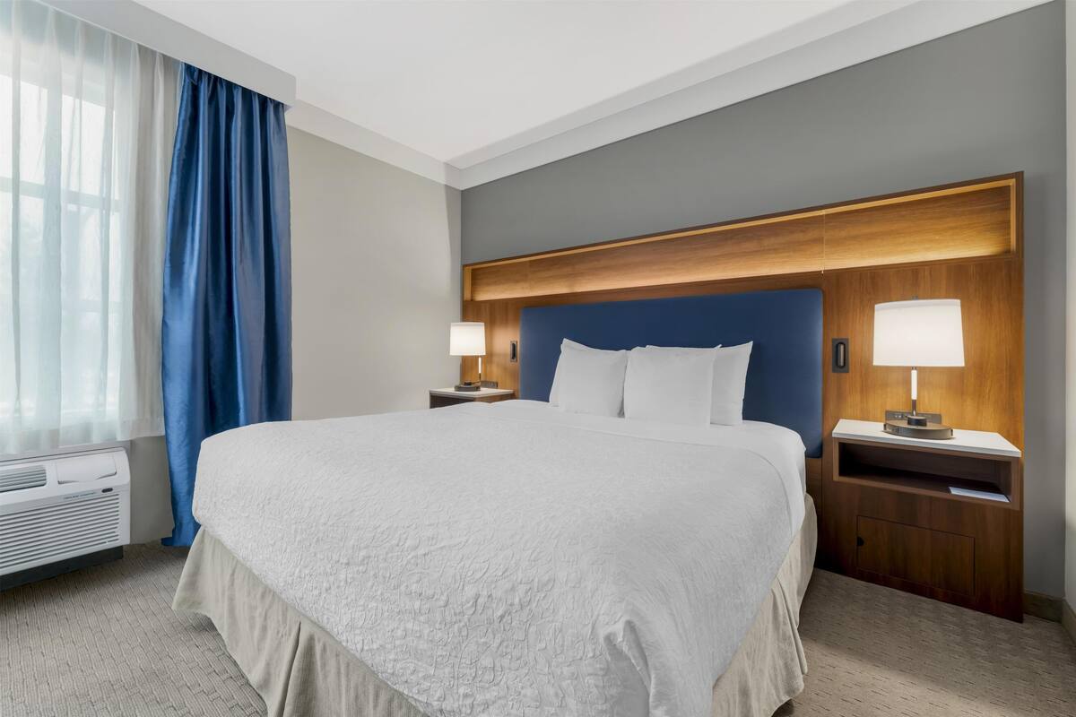 A hotel room with a large bed, a wooden headboard, two bedside tables with lamps, a window with blue curtains, and a wall-mounted air conditioner.