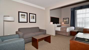 A hotel room with a grey sofa, a wooden coffee table, a desk with a laptop, a bed with a white comforter, and two framed pictures on the wall.
