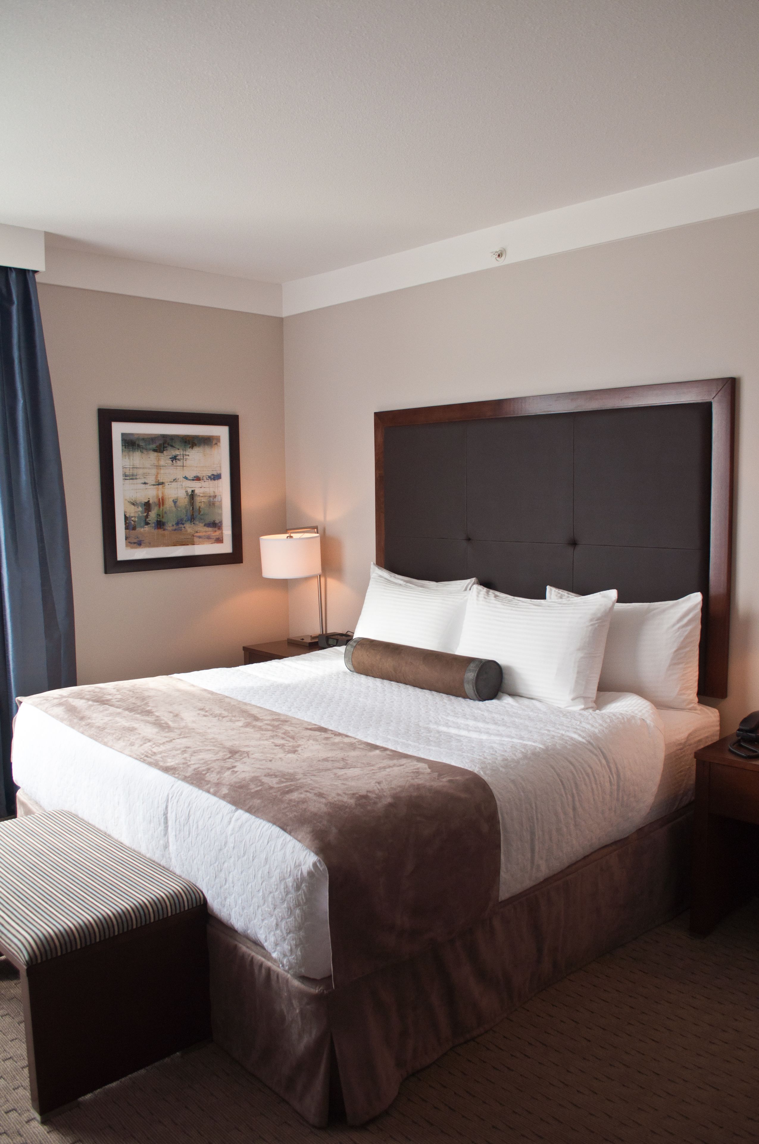 A hotel room with a large bed, a bedside table, a lamp, a framed picture on the wall, and a window with curtains.