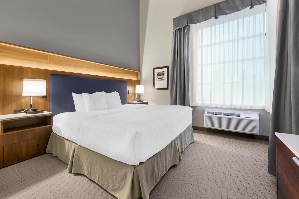 A hotel room with a large bed, bedside lamps, a wooden headboard, a window with curtains, and a wall-mounted picture.