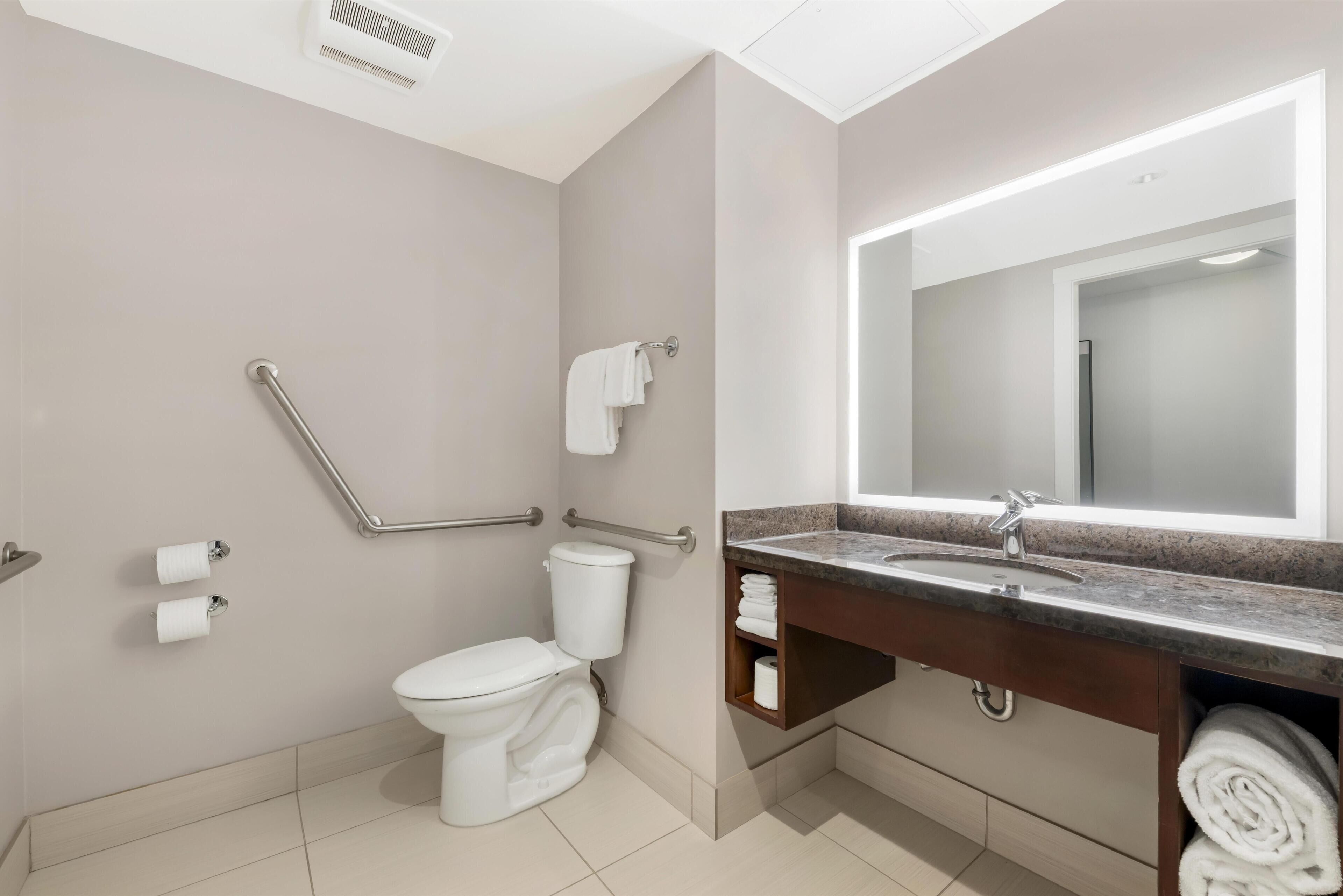 Un baño moderno con inodoro, barras de apoyo, lavamanos con espejo y toallero.
