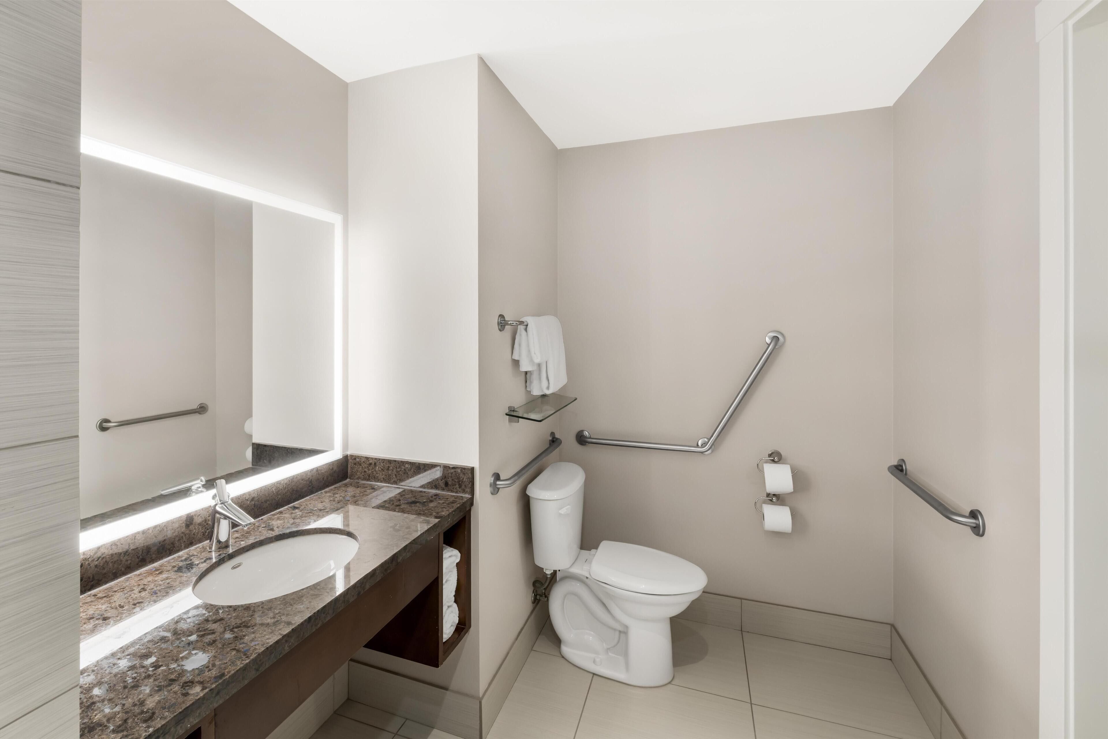 A modern bathroom with a granite countertop sink, a white toilet, and a walk-in shower with grab bars.