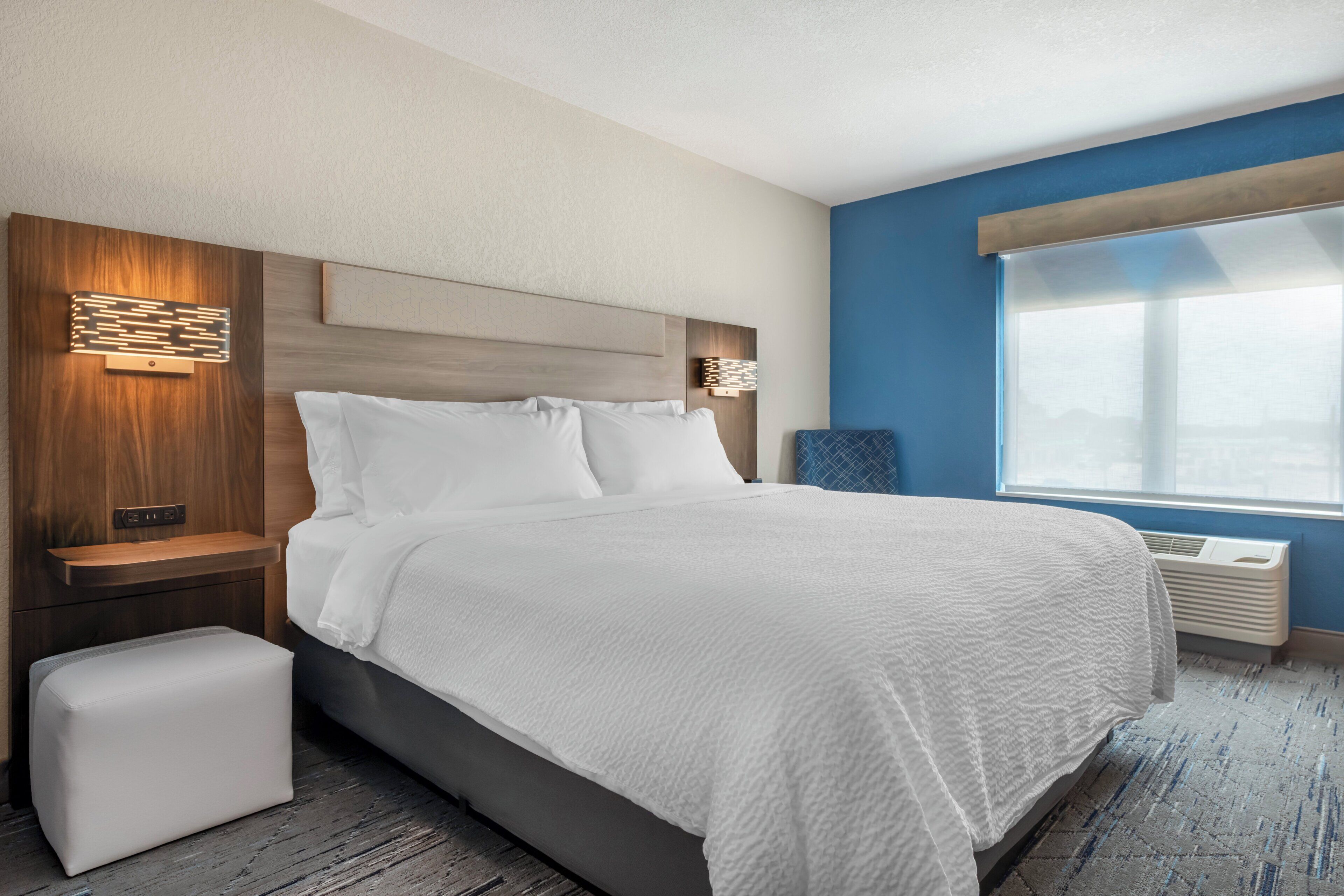 A hotel room with a large bed, bedside tables, a nightstand, a window with blinds, and a wall-mounted air conditioner.