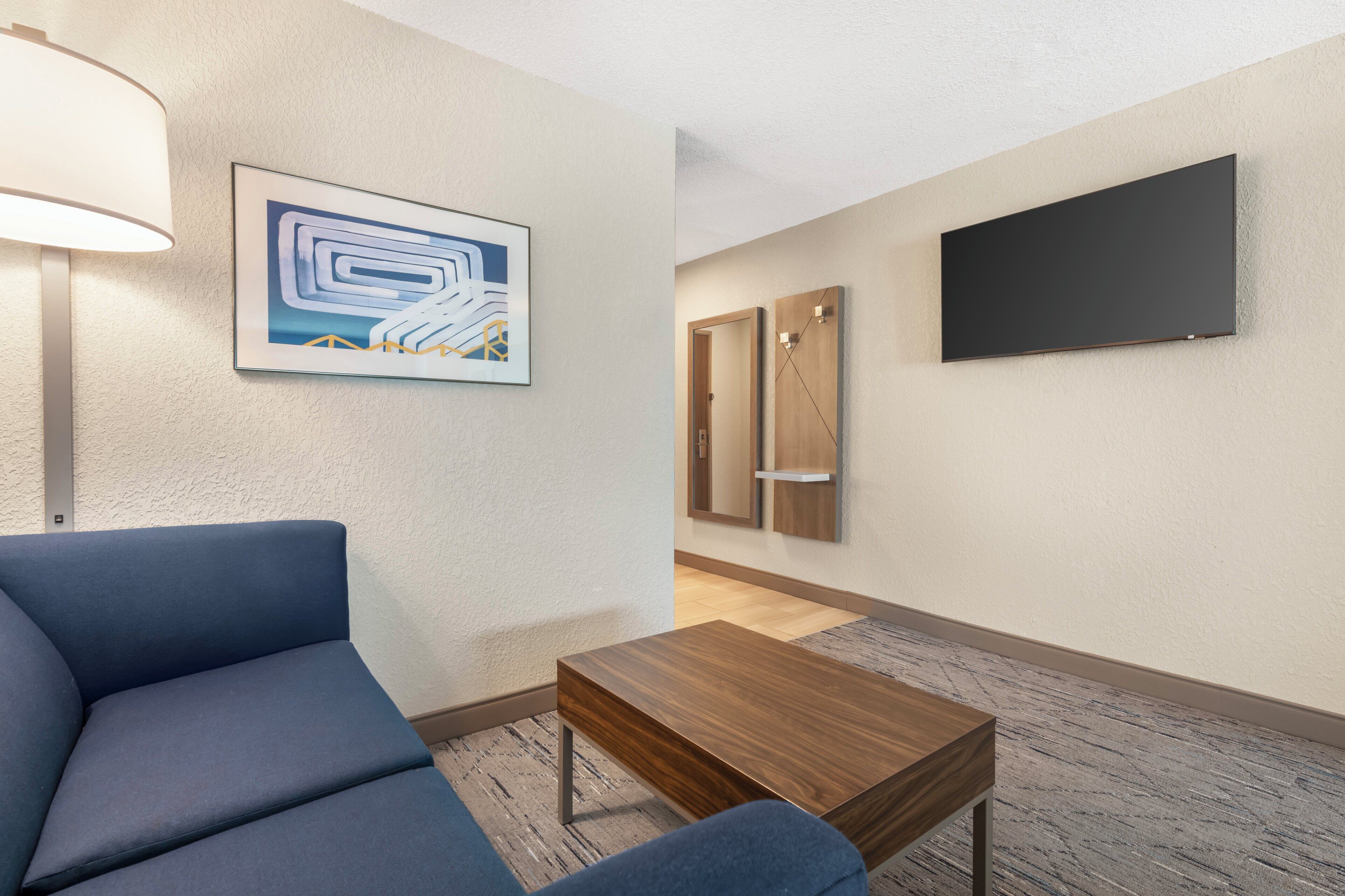 Ein Hotelzimmer mit einem blauen Sofa, einem Holztisch, einem Gemälde, einer Tür und einem Flachbildfernseher an der Wand.