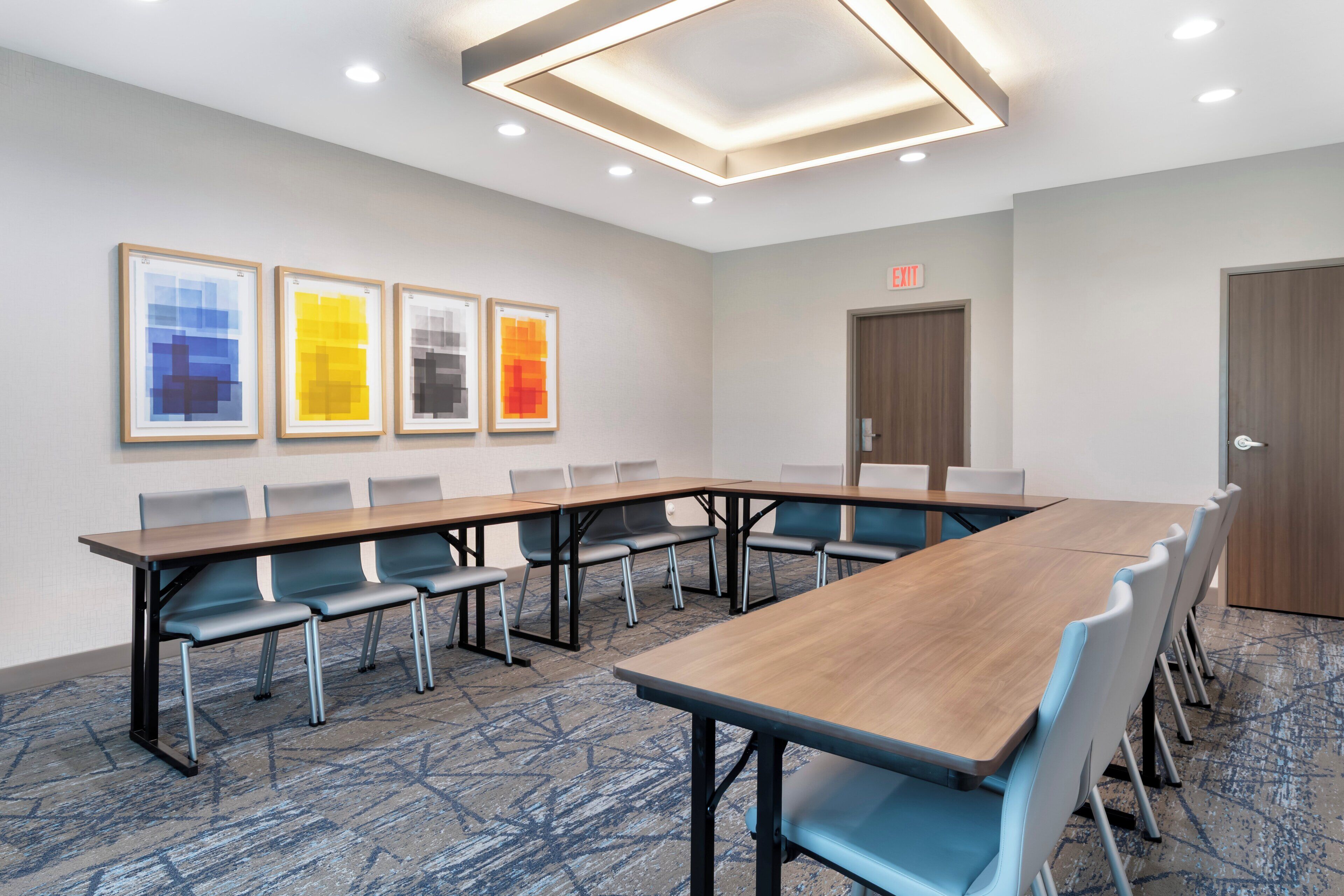 A conference room with a long table, chairs, framed artwork on the wall, and a door with an exit sign.