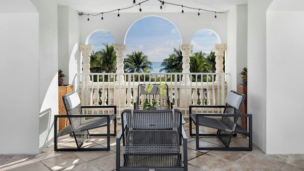 A balcony with three black chairs, a white railing, and a view of the ocean through semi-circular arches.