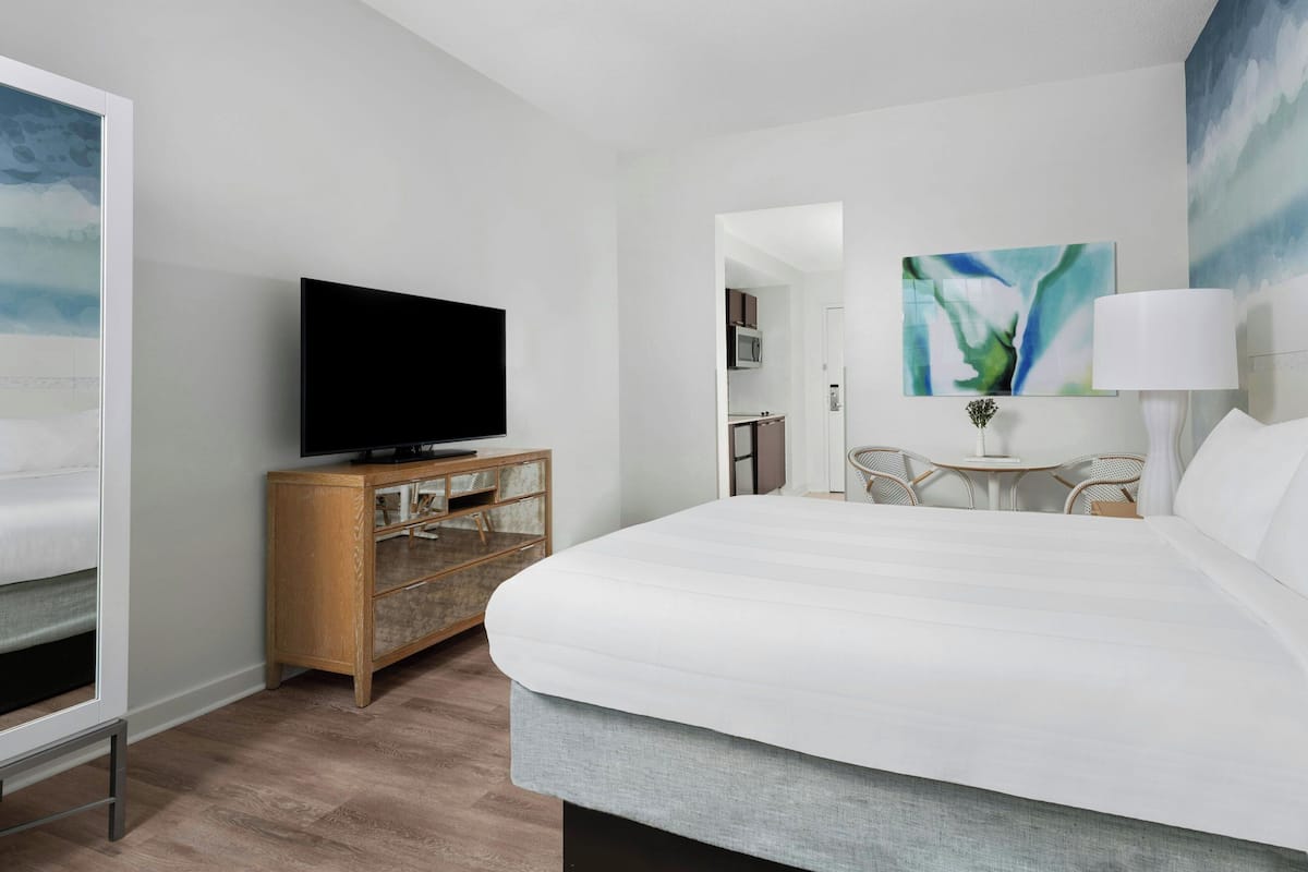 A hotel room with a large bed, a television on a wooden cabinet, a dining area with a table and chairs, and a kitchen in the background.