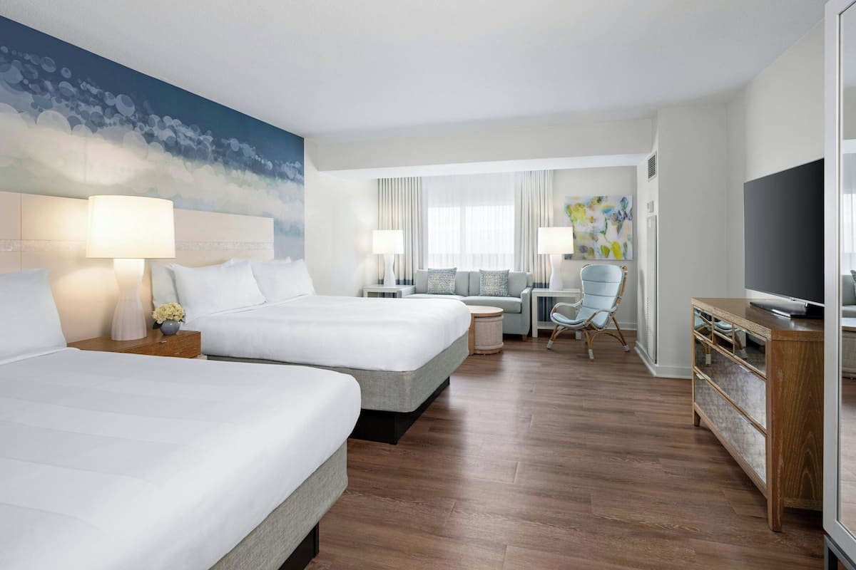 A hotel room with two beds, a desk, a chair, a TV, and a painting on the wall.