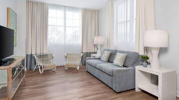 A modern living room with a grey sofa, two wicker chairs, a white side table with a lamp, and a TV on a wooden console.