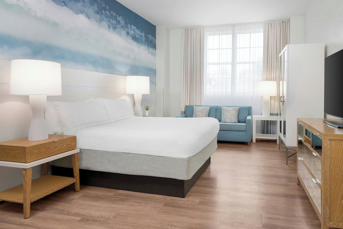A hotel room with a large bed, two bedside tables with lamps, a TV, a sofa, and a large window with curtains.
