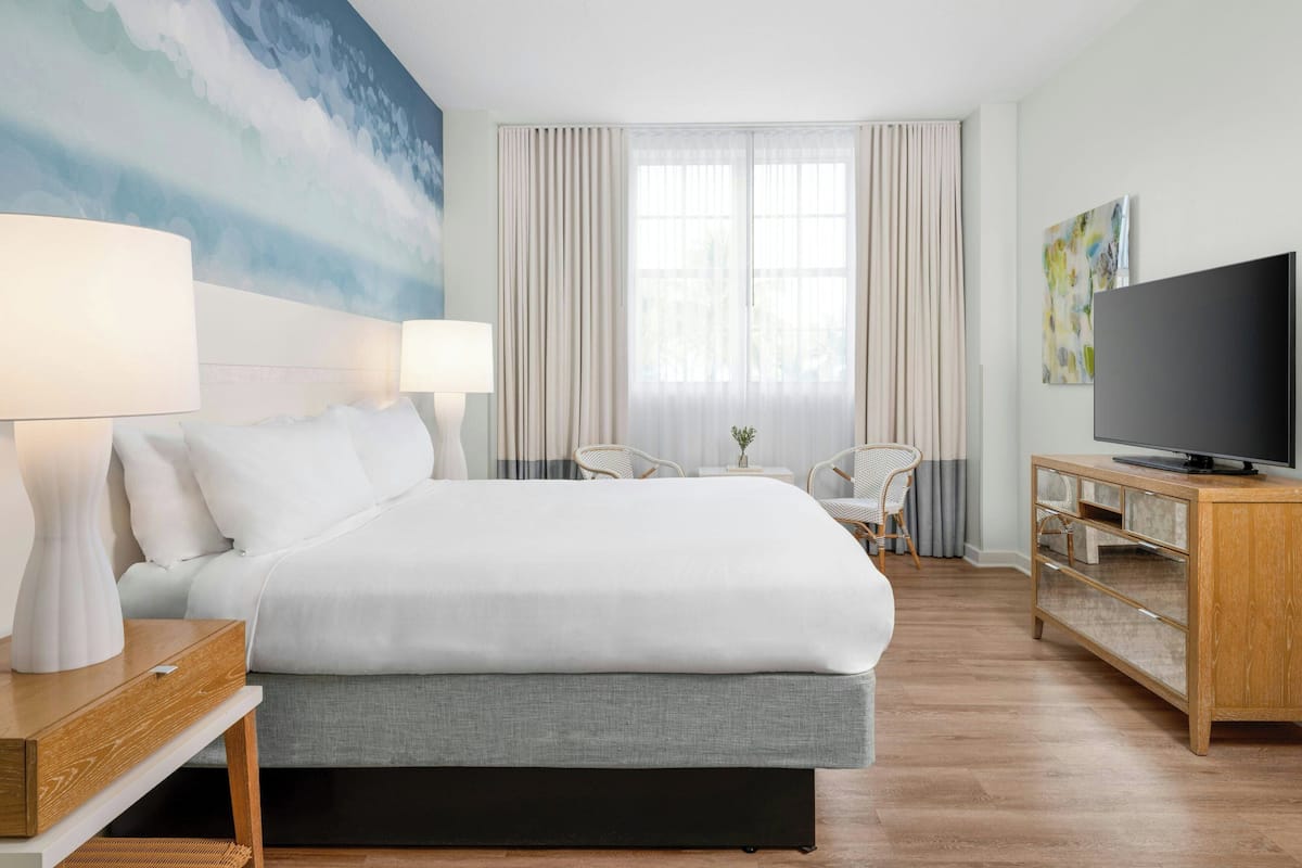 A hotel room with a large bed, two bedside lamps, a television on a wooden cabinet, a painting on the wall, and a window with curtains.