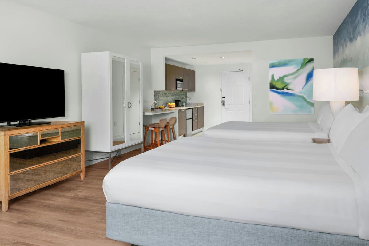 A modern hotel room with a large bed, a TV, a kitchenette, and a painting on the wall.