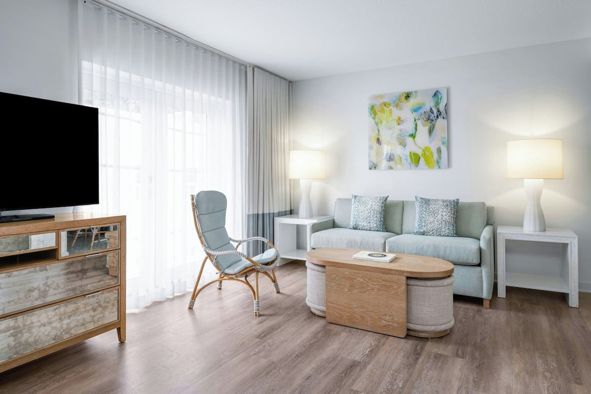 A modern living room with a flat-screen TV, wicker chair, two side tables with lamps, a sofa with patterned cushions, and a painting on the wall.