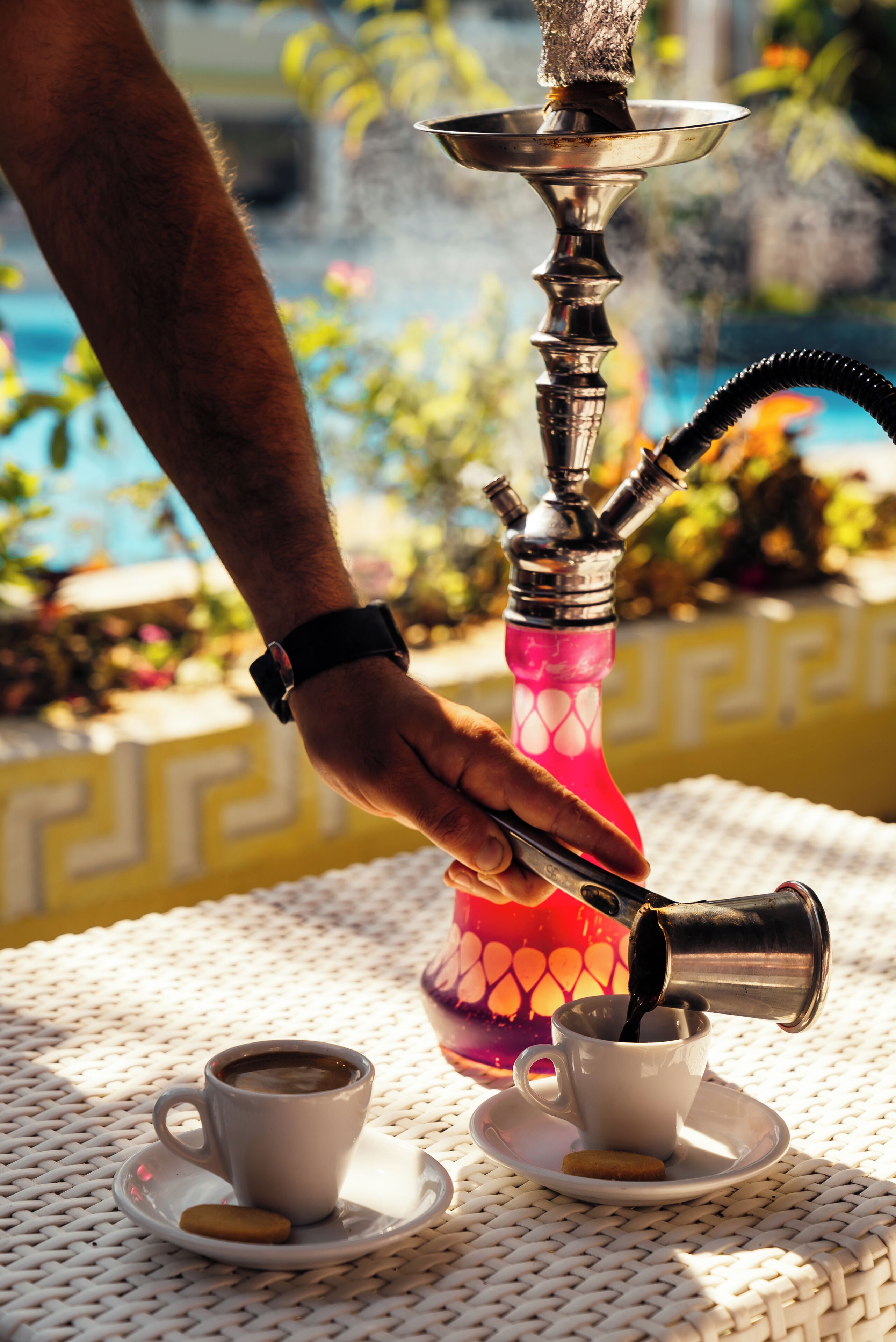 A hookah with a pink base, a cup of coffee, and a plate with a cookie on a white woven table.