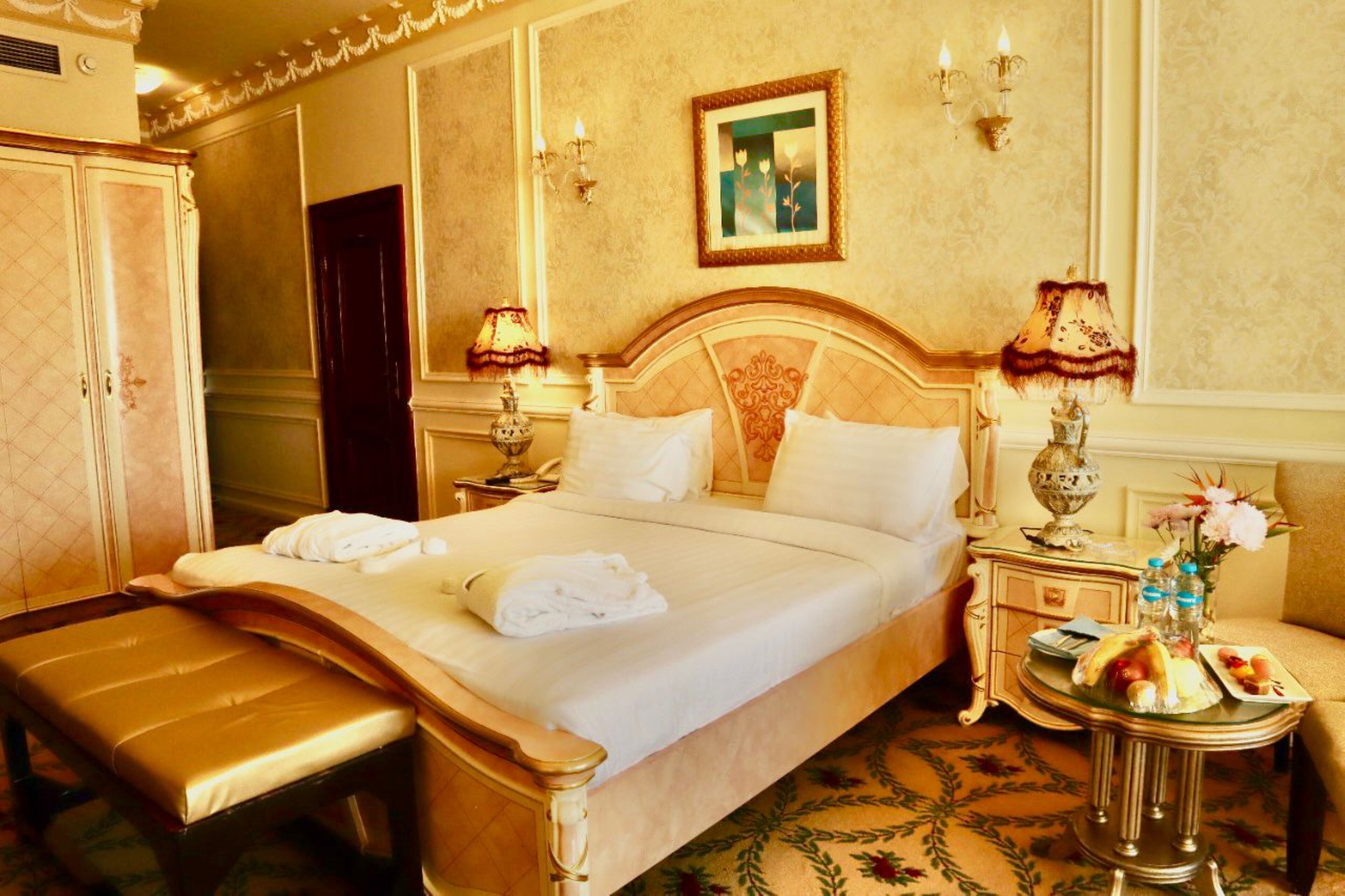Ein Hotelzimmer mit einem großen Bett, zwei Nachttischlampen, einem kleinen Tisch mit Blumenvase und einer Bank.