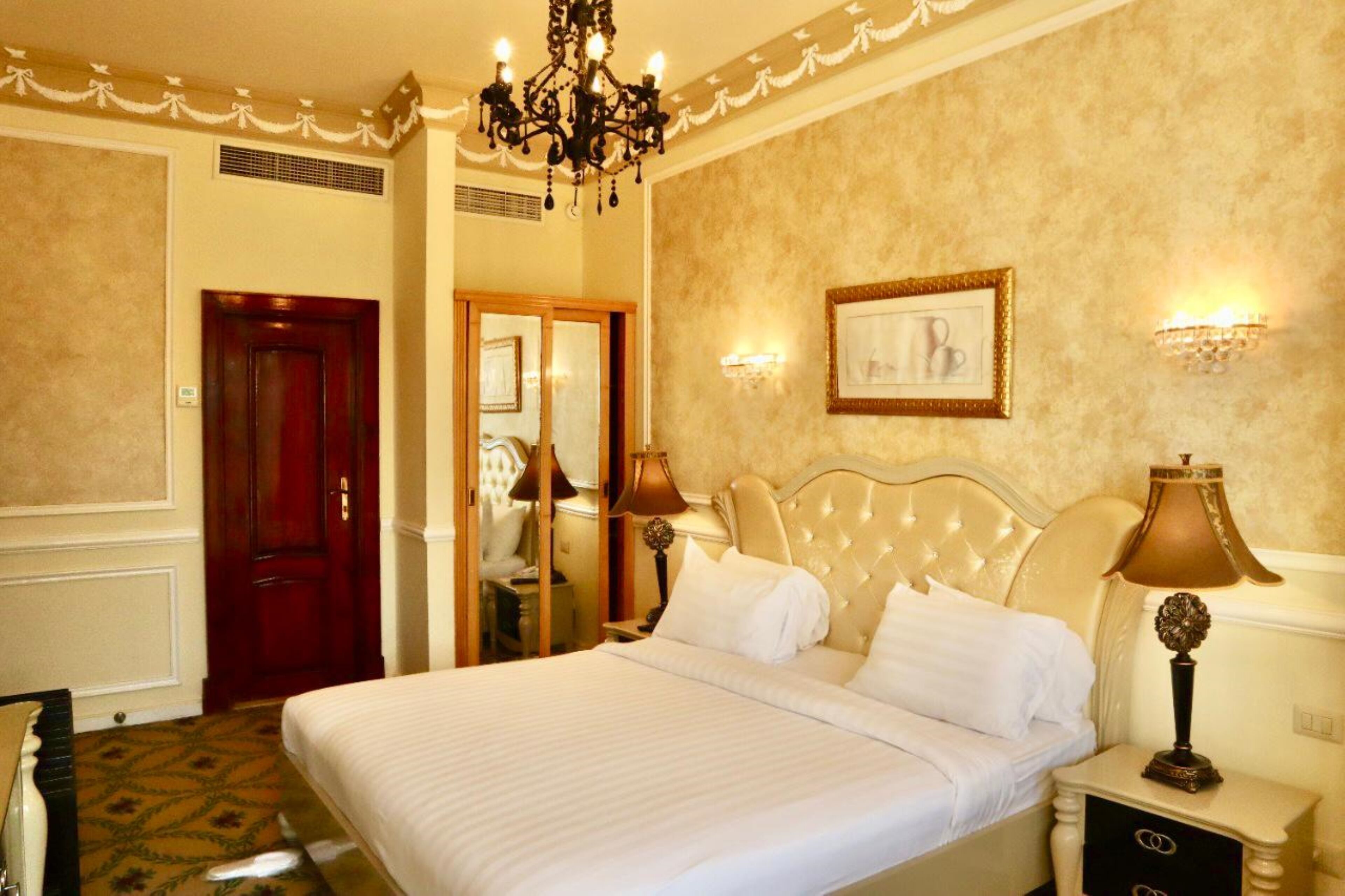 A hotel room with a large bed, a chandelier, a framed picture on the wall, and a nightstand with a lamp.