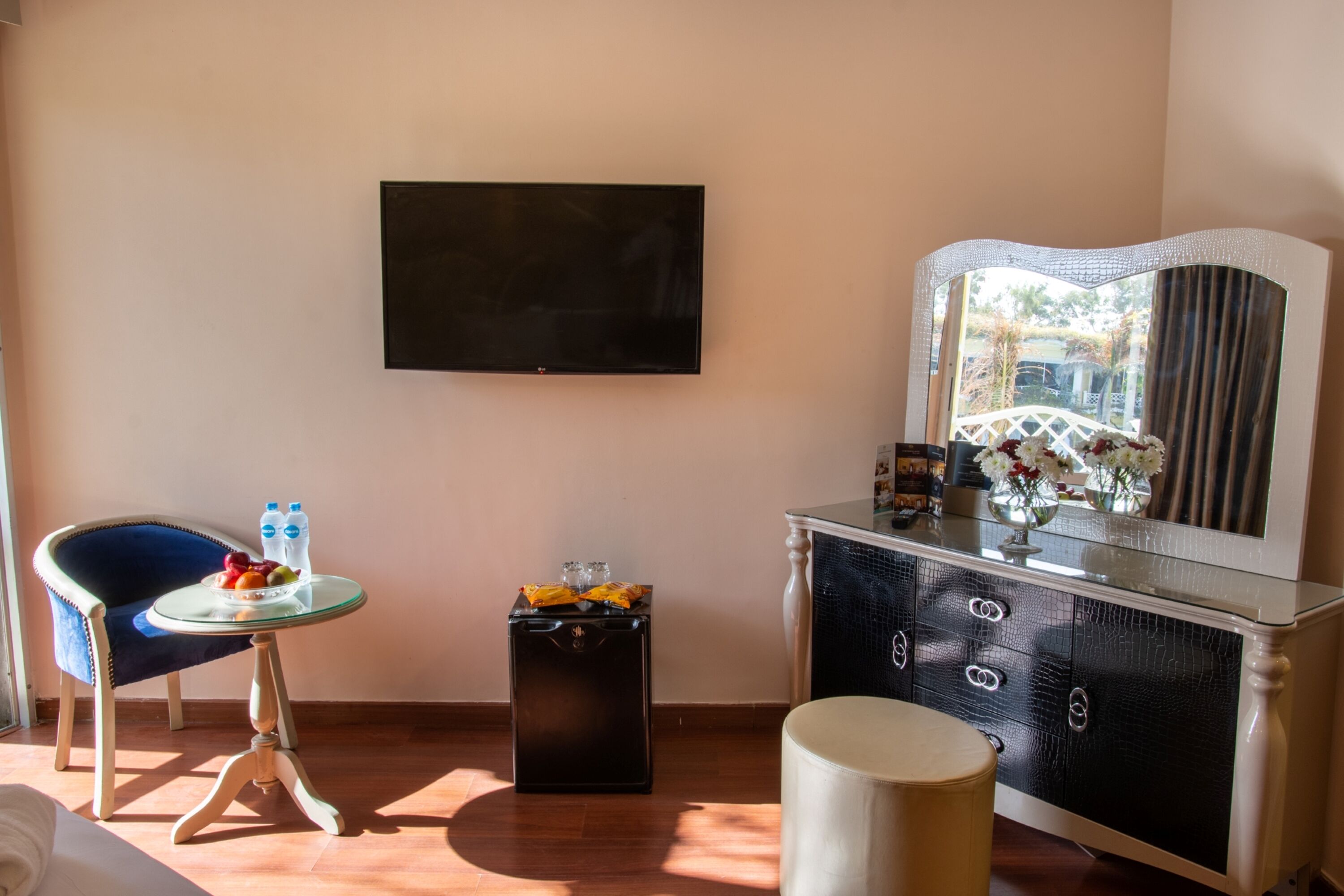 A room with a TV mounted on the wall, a small round table with a chair, a cabinet with drawers, and a large mirror.
