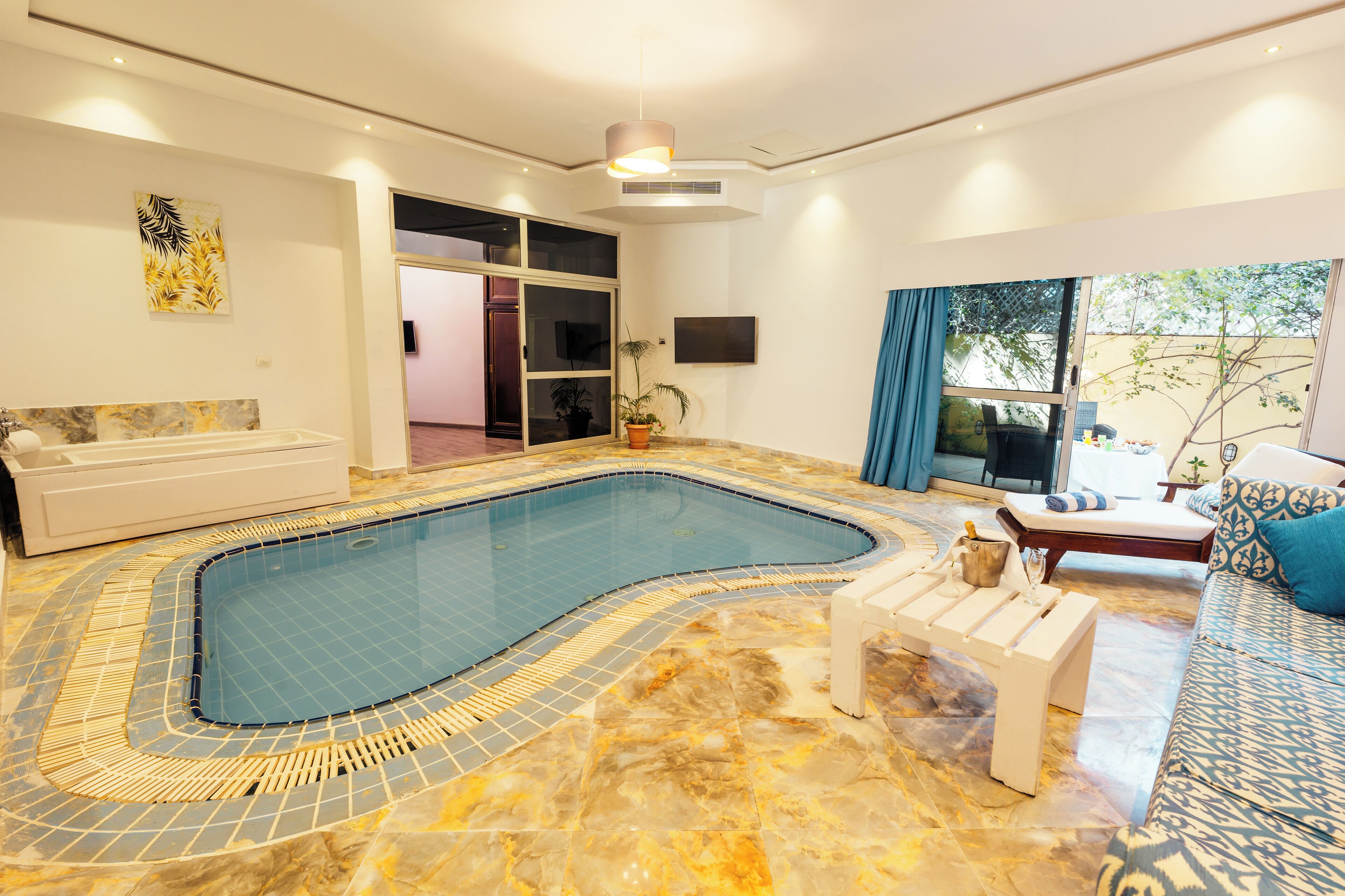 En rummelig indendørs swimmingpool med et bølgende design, et siddeområde med sofa og lænestole, samt et tv ophængt på væggen.