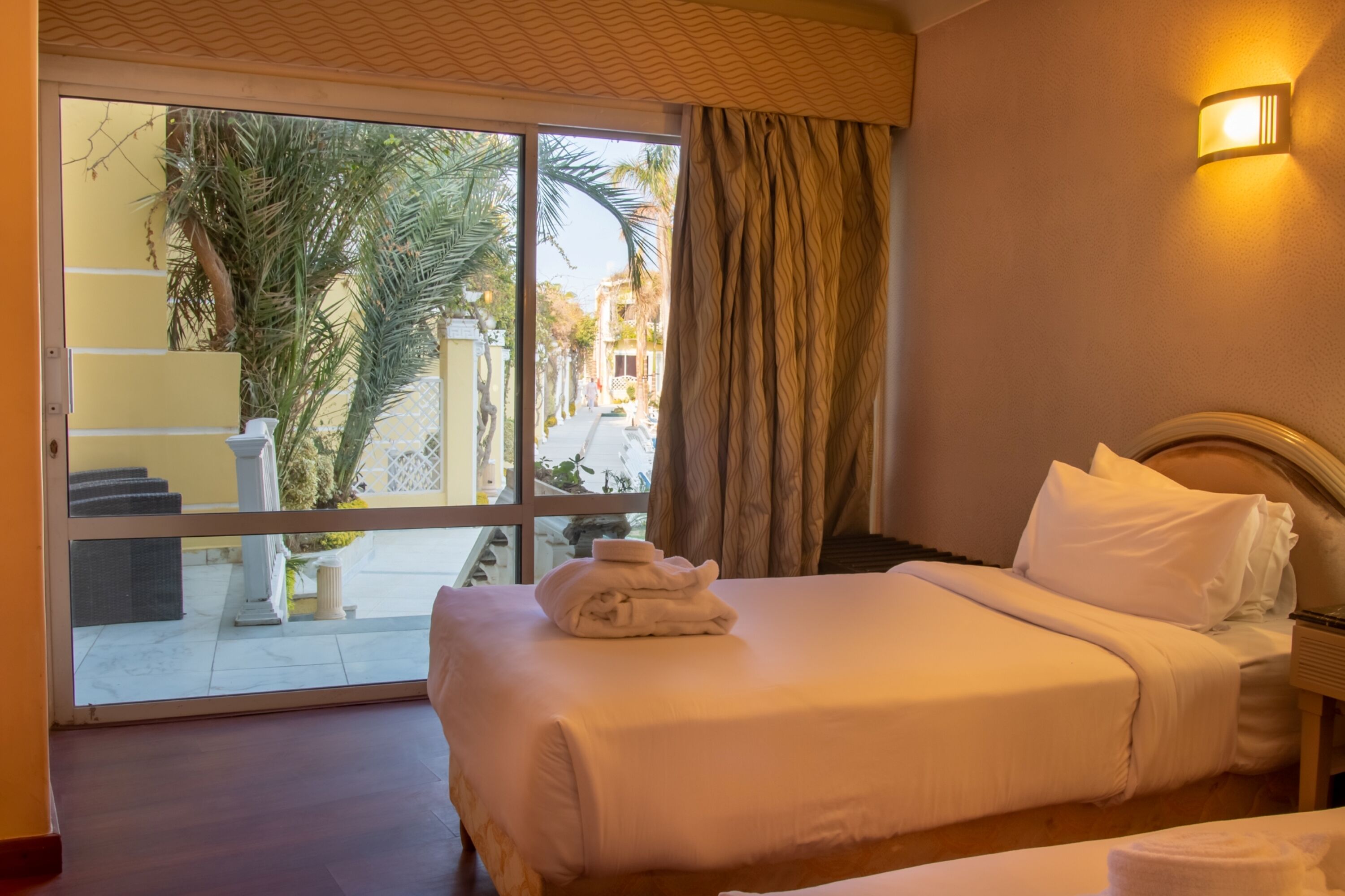 A hotel room with a large sliding glass door leading to a balcony, a bed with white linens, a nightstand, and a wall-mounted light.