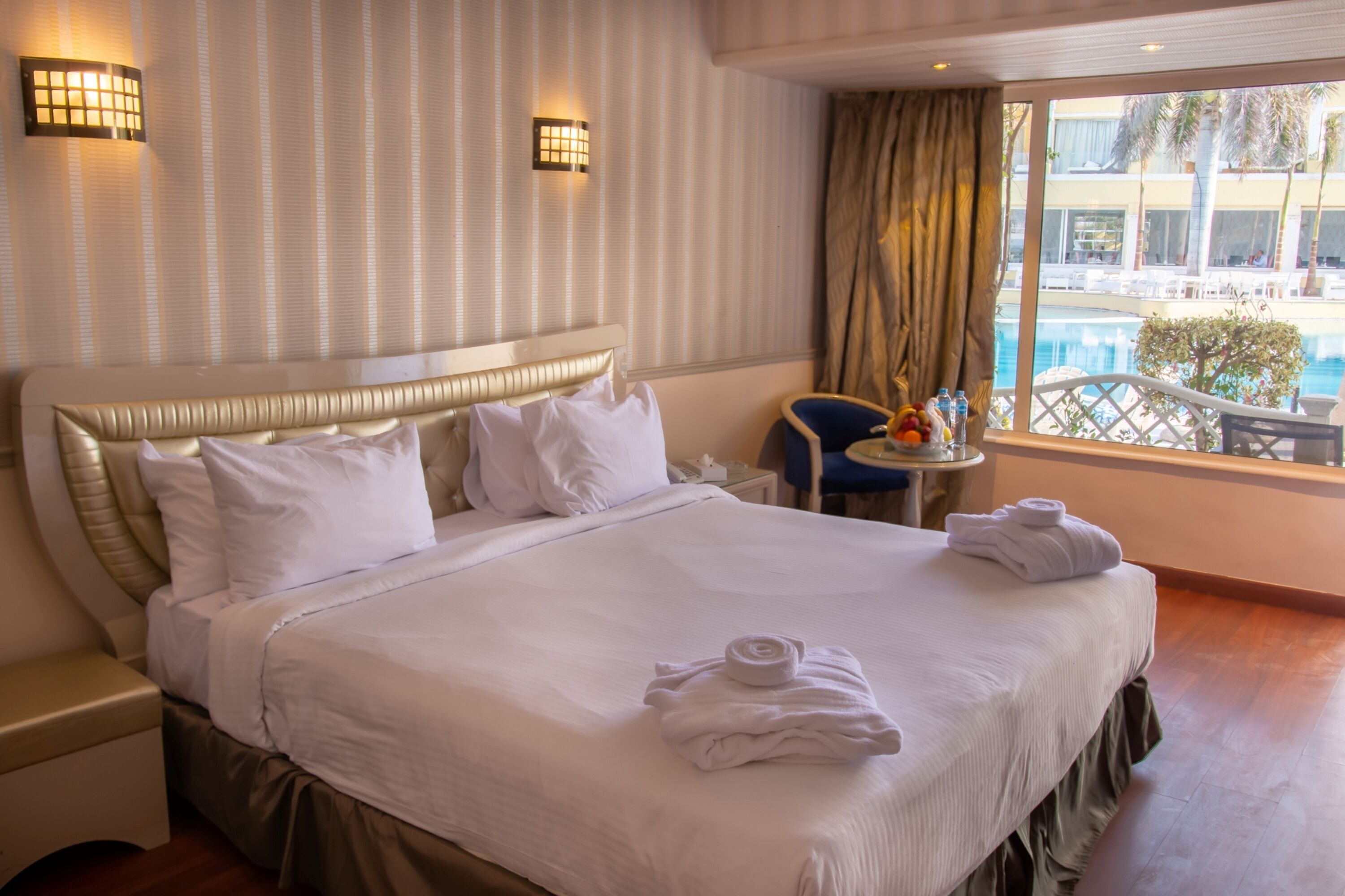 A hotel room with a large bed, two bedside tables, a window with a view of a pool and palm trees, and two wall-mounted lights.