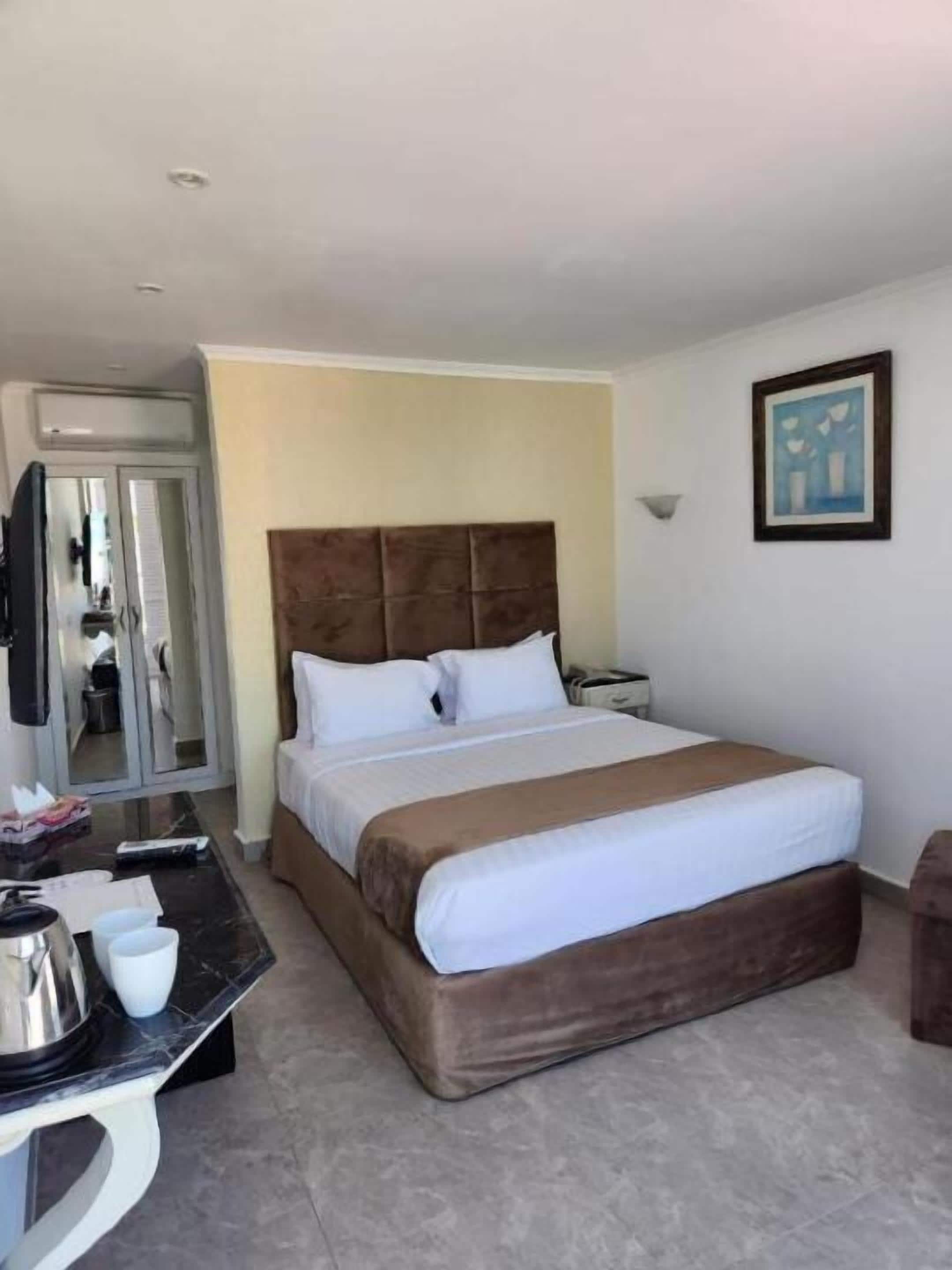 A hotel room with a large bed, a desk with a kettle and cups, a television, and a framed picture on the wall.
