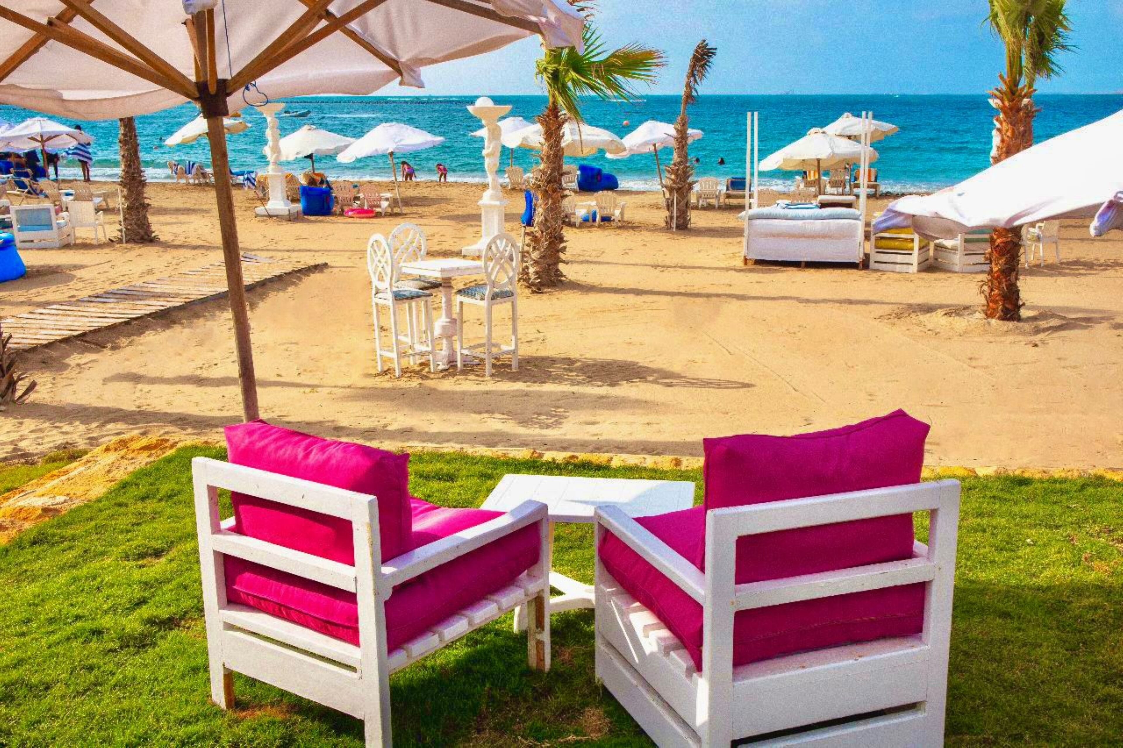 Un entorno frente al mar con sillas de descanso blancas, una mesa, palmeras y sombrillas.