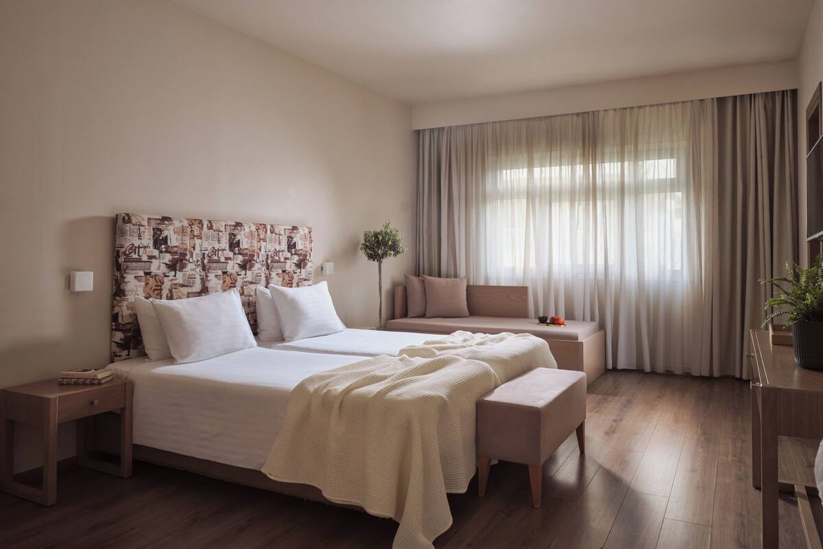 A modern hotel room with a large bed, a bedside table, a bench, a sofa, and a large window with curtains.