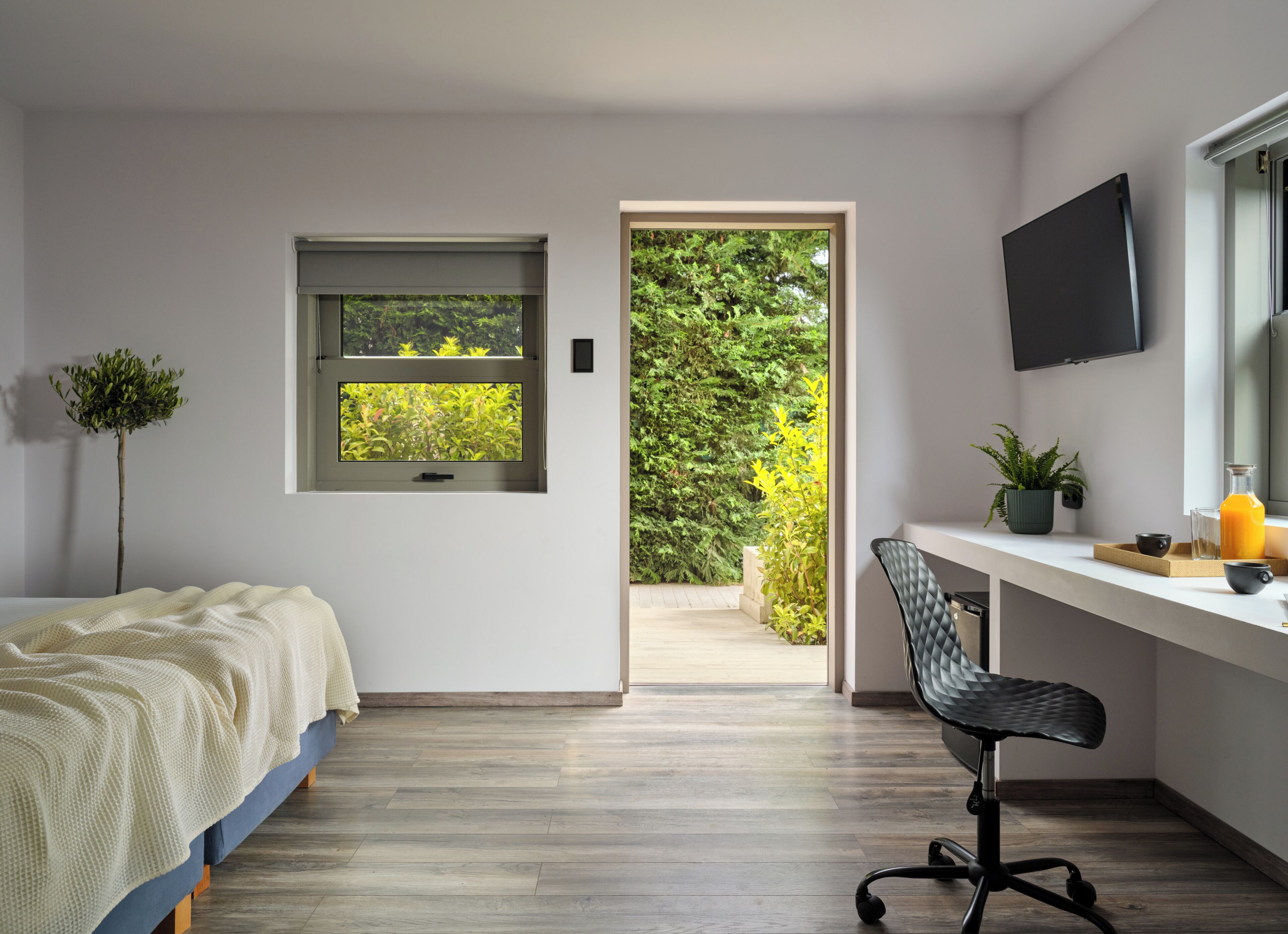 Un dormitorio con cama, escritorio, silla, televisor, ventana y puerta que da a un balcón con plantas.