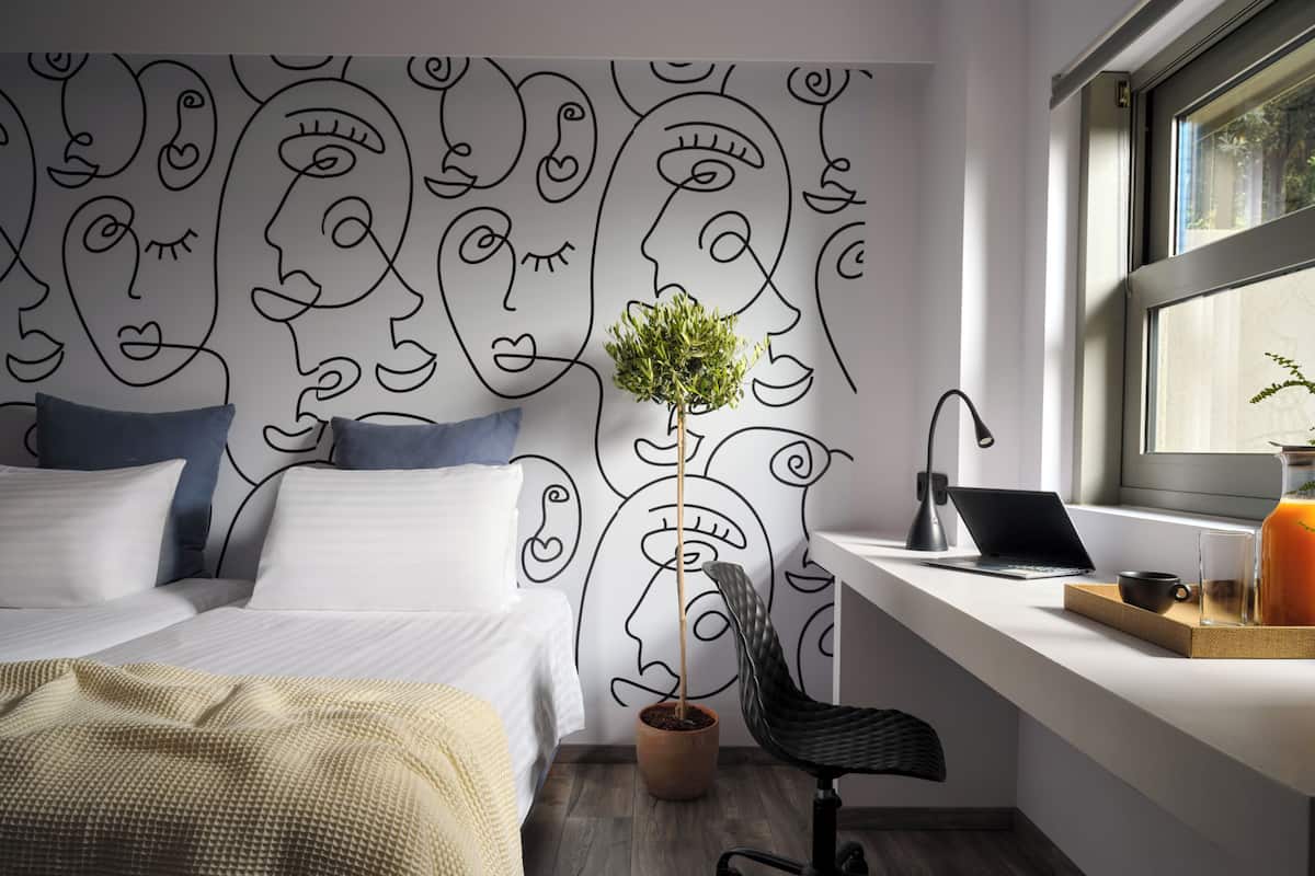 A modern bedroom with a bed, a desk, a laptop, a potted plant, and a window.