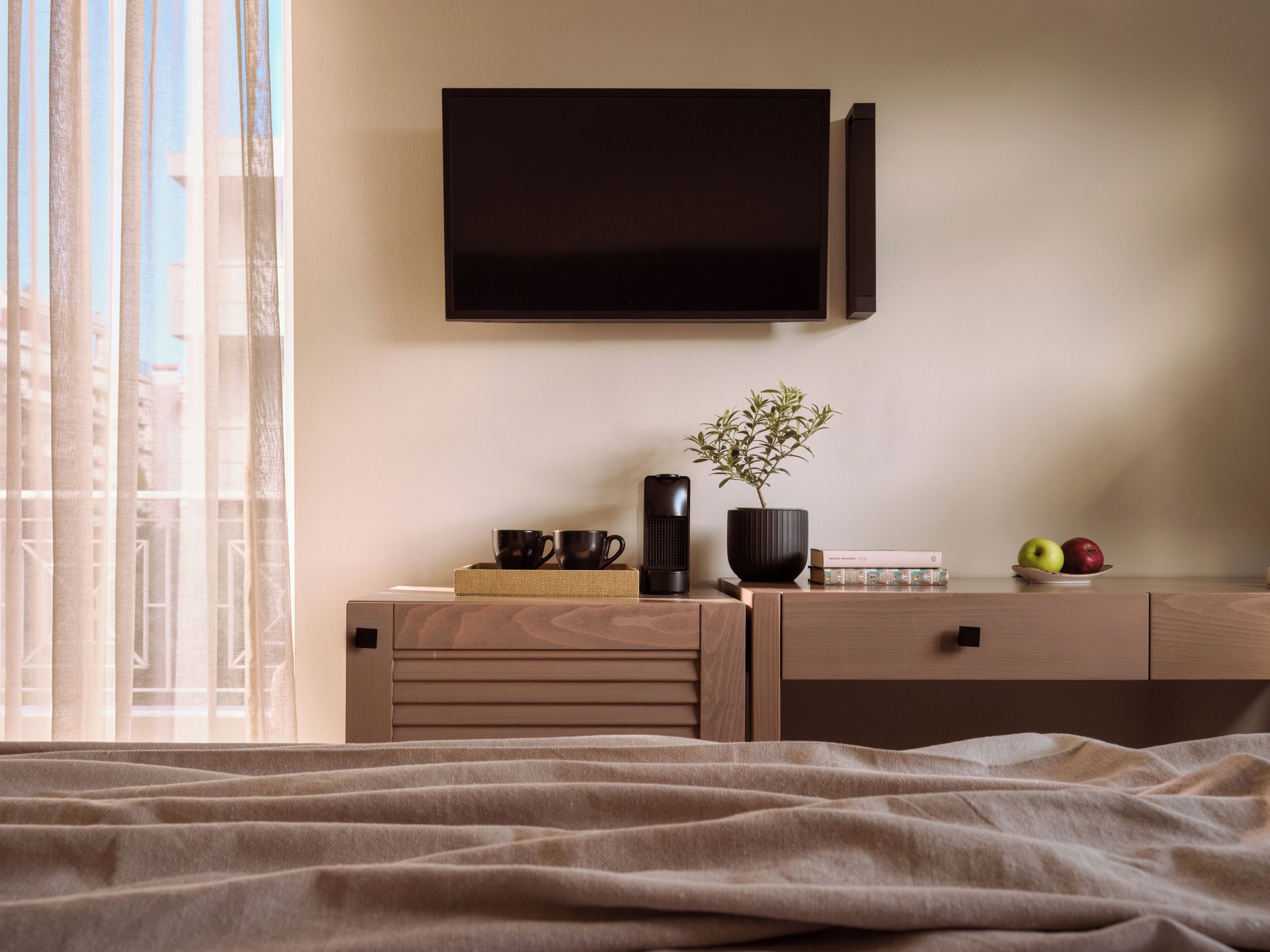 A bedroom with a bed, a bedside table with a plant, a speaker, and a flat-screen TV mounted on the wall.