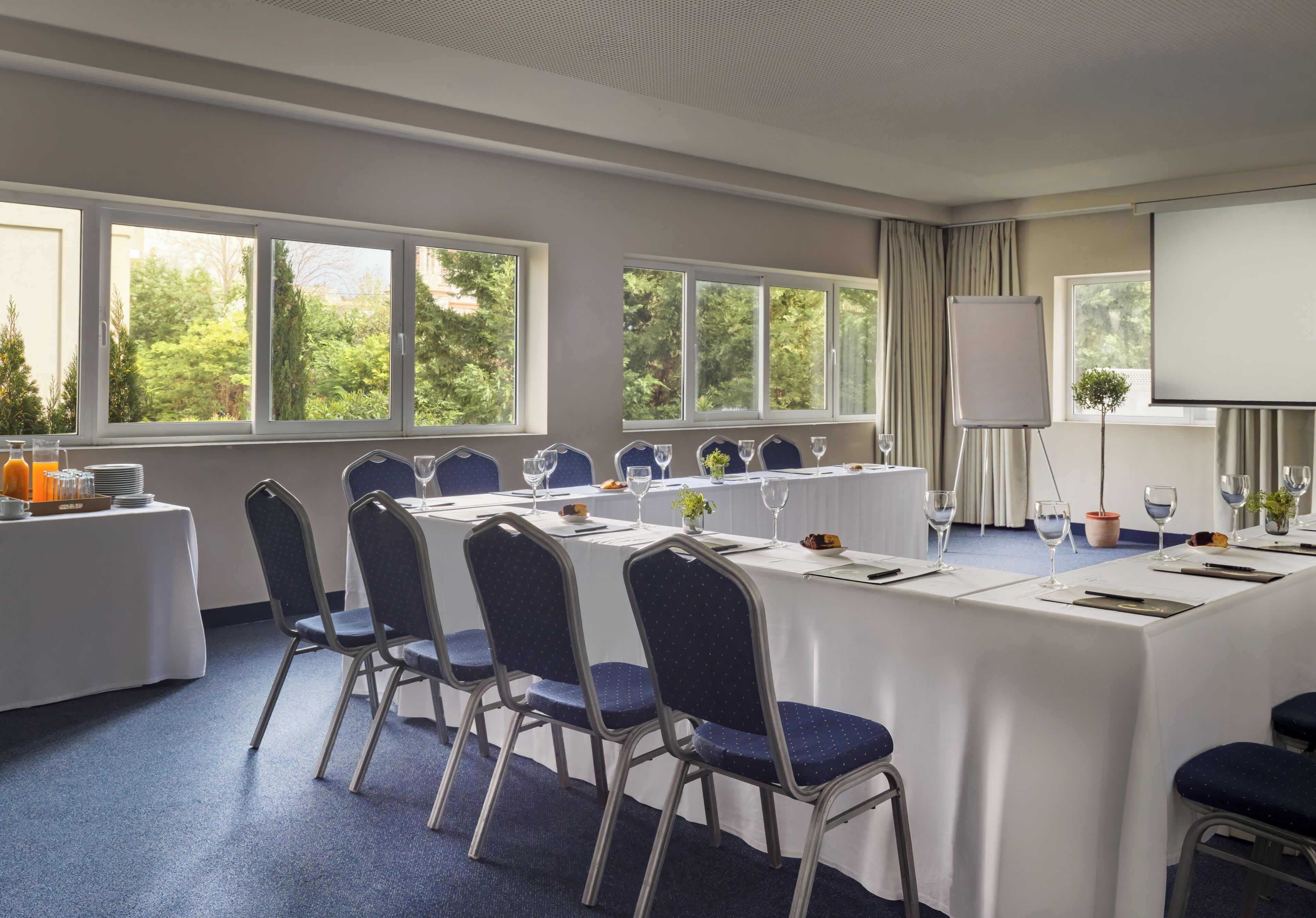 Uma sala de conferências com uma mesa comprida, cadeiras, uma tela de projetor e janelas com vista para árvores.