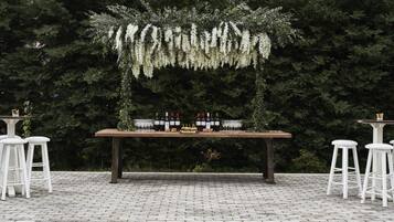 Un área de estar al aire libre con una mesa de madera dispuesta para una comida, rodeada de taburetes blancos, frente a un fondo de follaje verde denso.