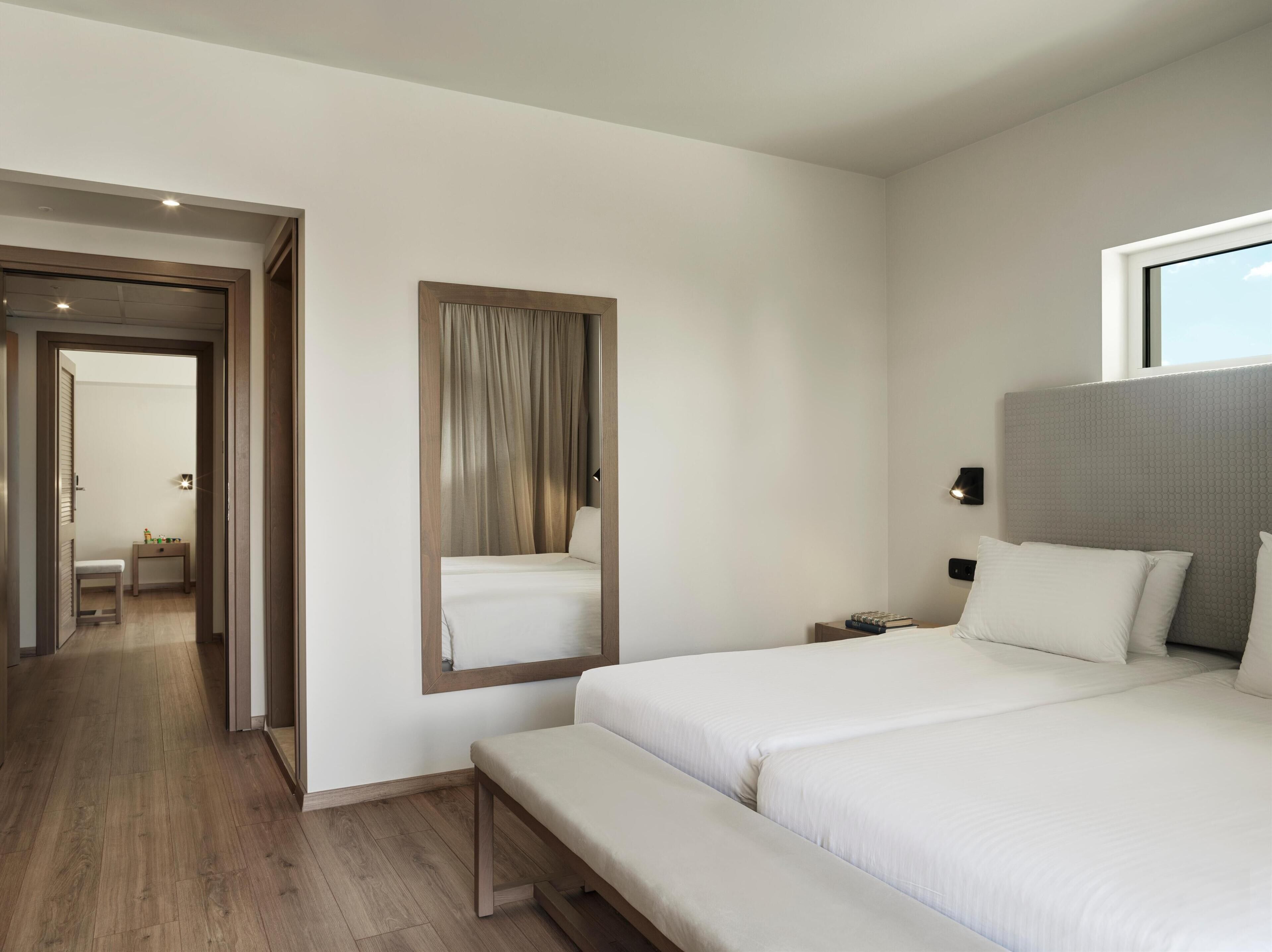 Quarto de hotel moderno com uma cama grande, uma mesinha de cabeceira, um espelho e vista para o corredor.
