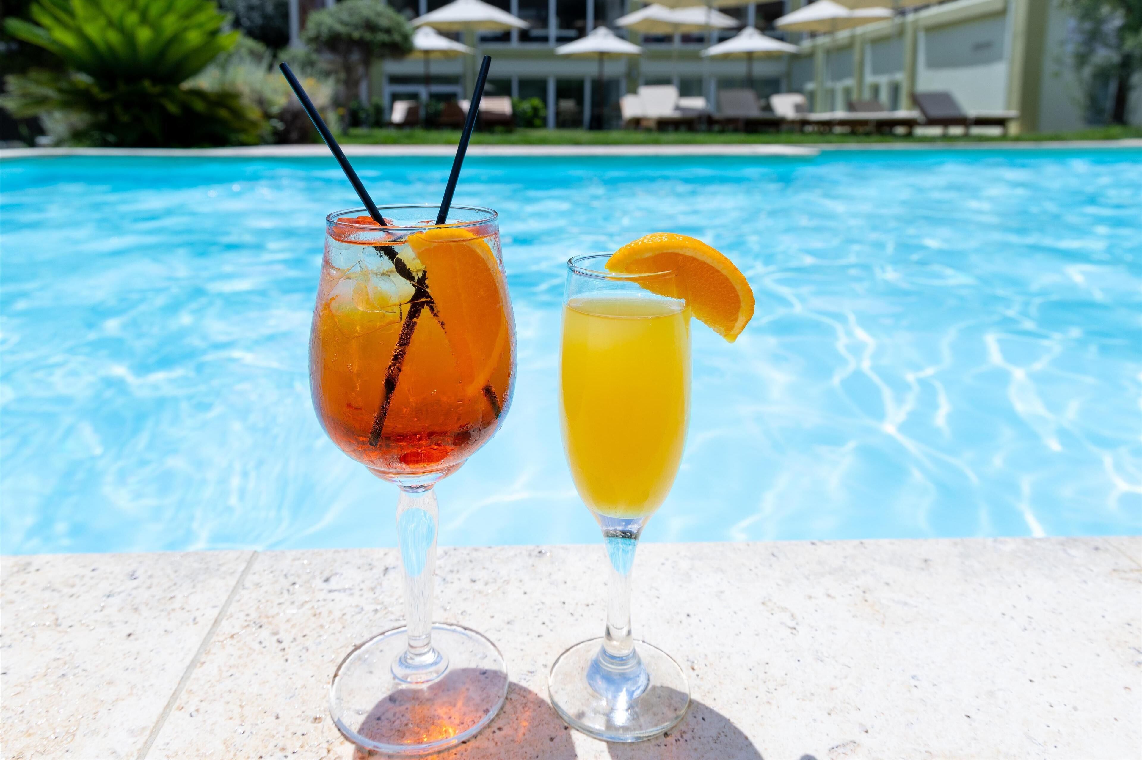 Two drinks on a poolside table with a pool and lounge chairs in the background.