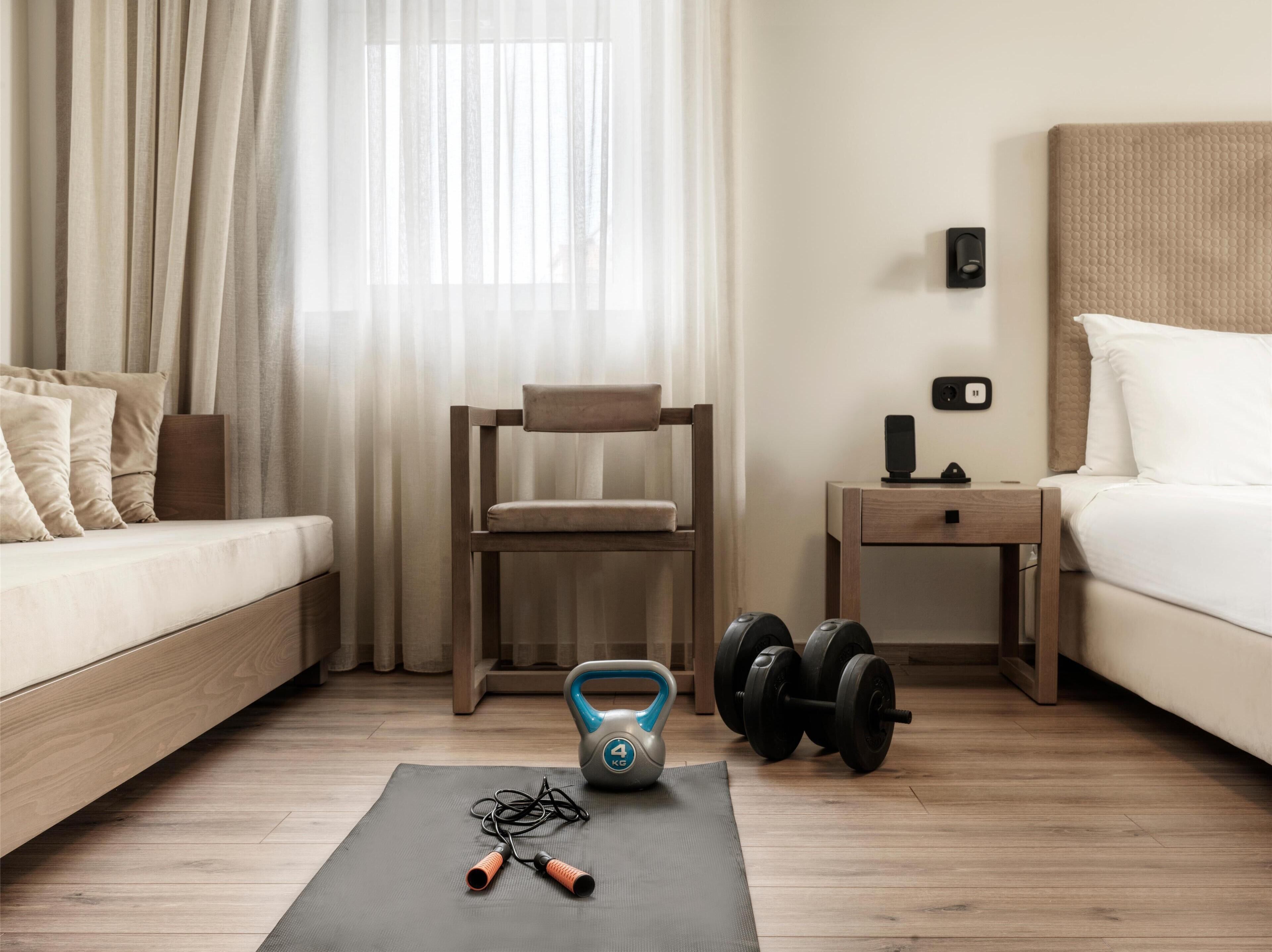 A modern hotel room with a wooden floor, a bed, a desk, and a chair. There are dumbbells and a jump rope on the floor.