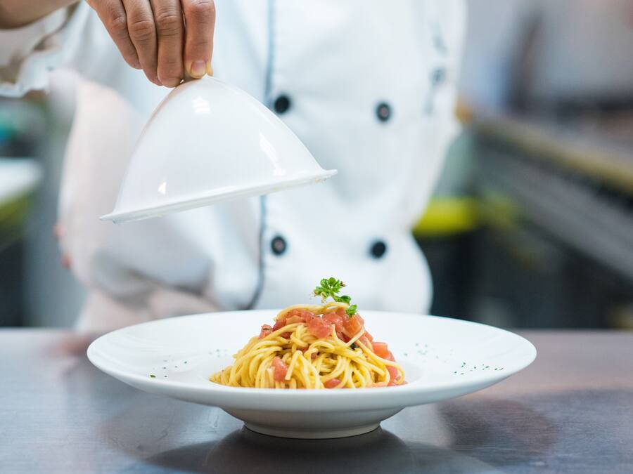 A chef holding a white dome over a plate of spaghetti with a garnish.