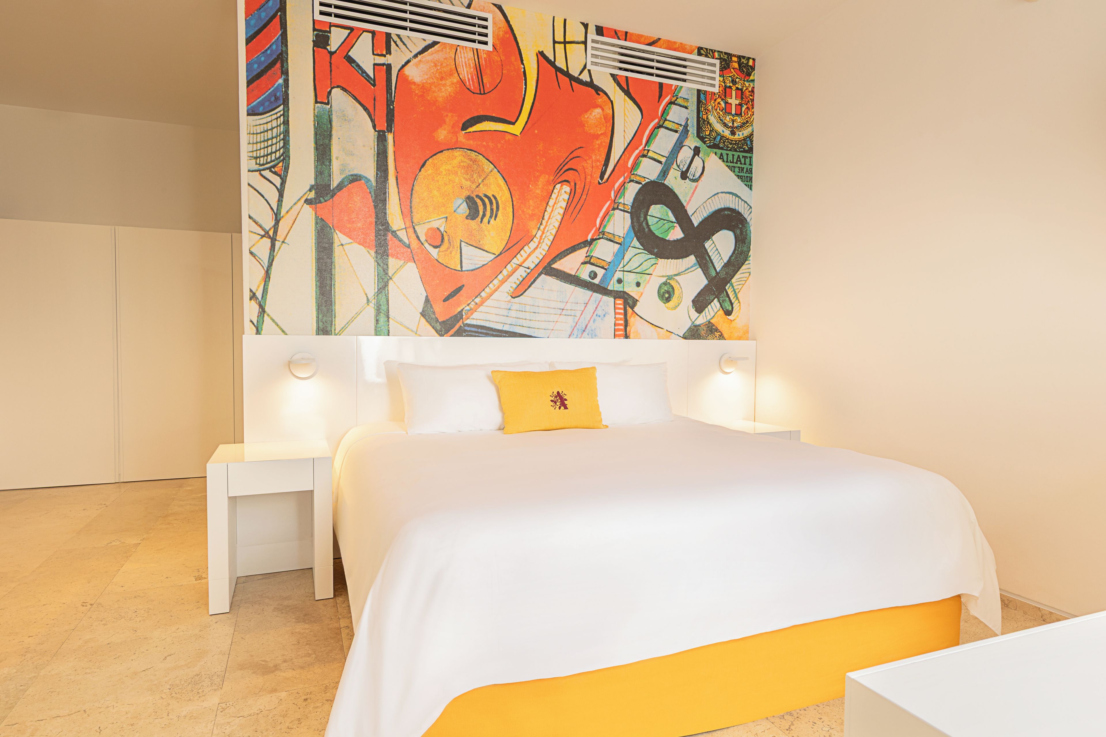 Habitación de hotel moderna con un cuadro abstracto grande en la pared, una cama blanca con un detalle amarillo y una mesita de noche pequeña.
