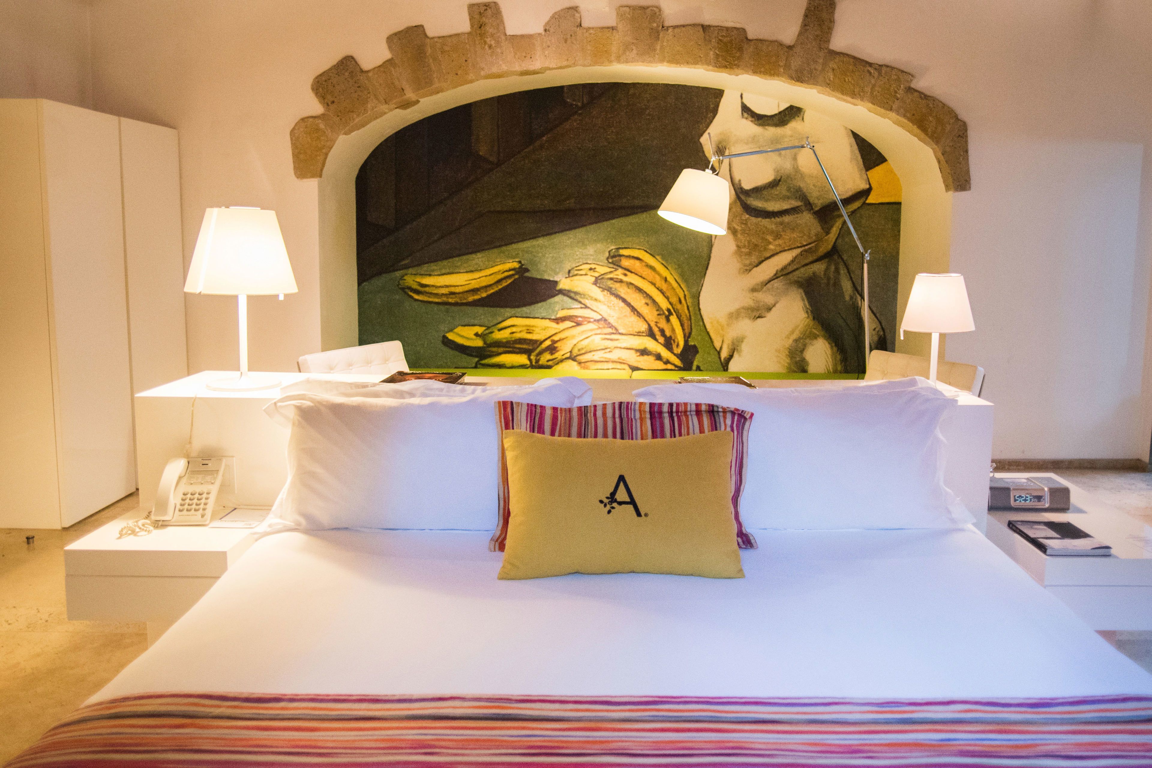 Habitación de hotel con cama, dos lámparas de noche, un teléfono, un cuadro de plátanos y un almohadón con la letra 'A'.