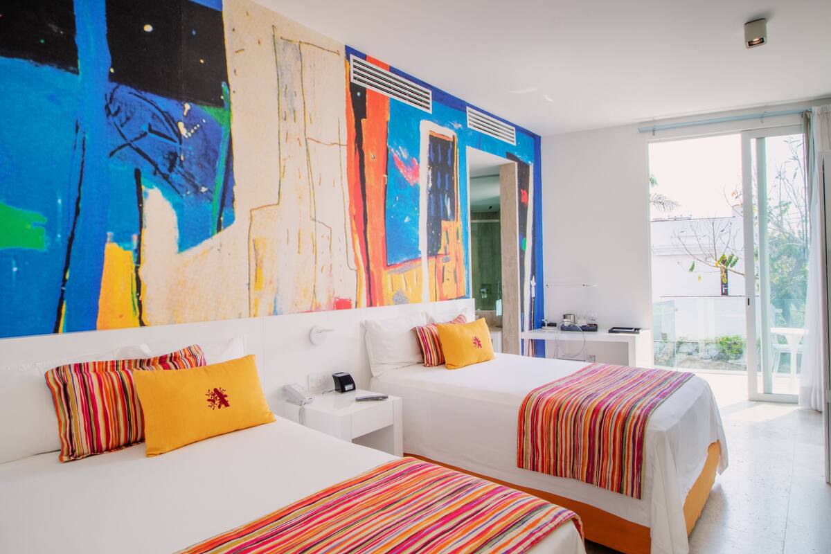A hotel room with two beds, a large abstract painting on the wall, a balcony with a view, and a desk with a phone and a coffee maker.