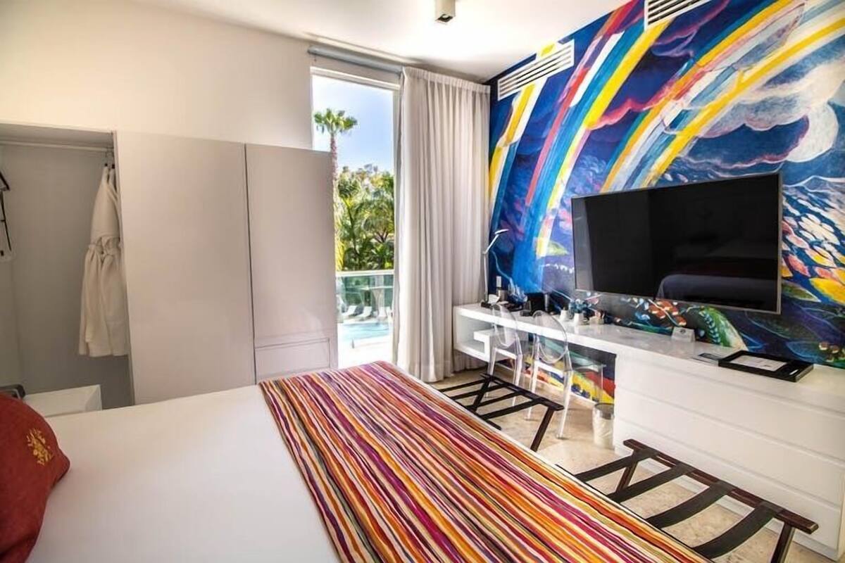 A hotel room with a bed, a desk, a TV, and a colorful abstract painting on the wall.